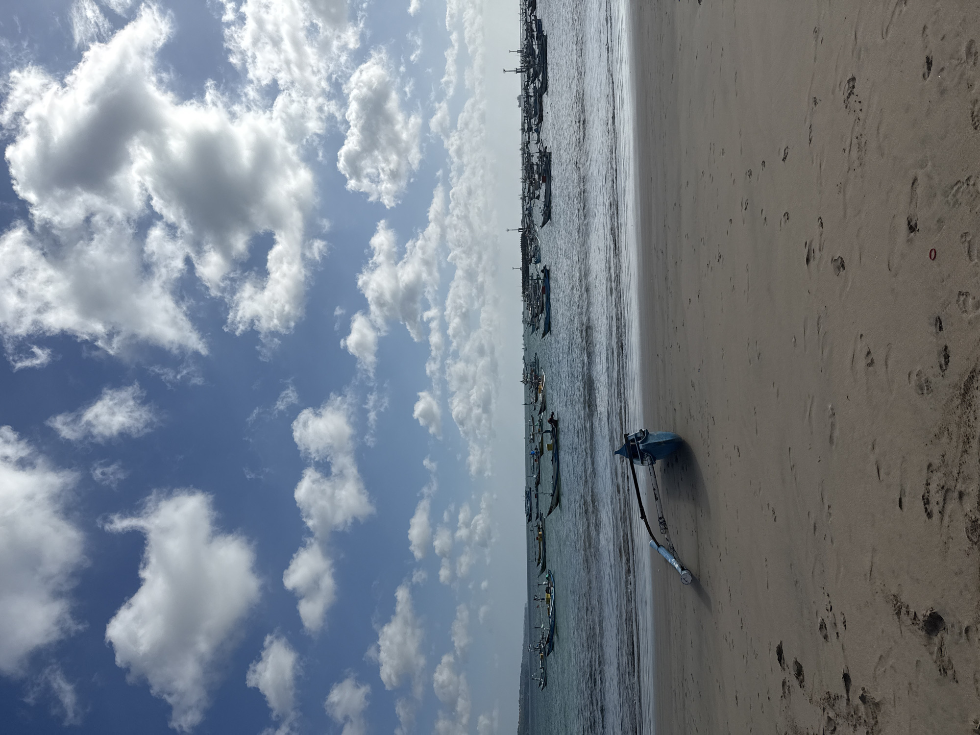 The photograph captures a beach scene in Kuta, Indonesia, under a partly cloudy sky. Numerous boats are anchored in the ocean near the shore. On the sandy beach, a blue object with metallic components rests, possibly a piece of equipment or gear. The sand is marked with footprints.
