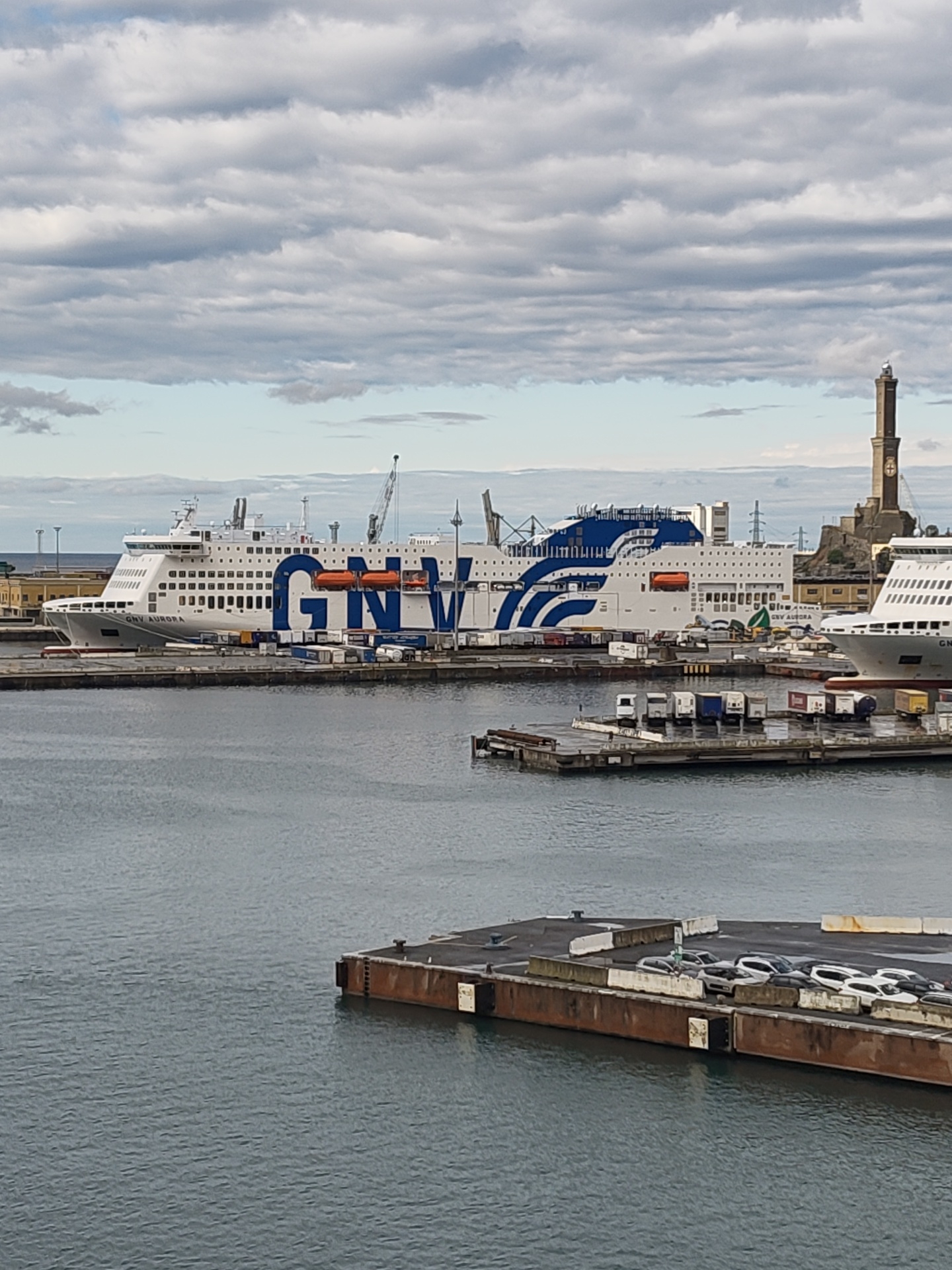 The image depicts a harbor scene, prominently featuring two large GNV ferries docked alongside a pier. The ships are painted white with large, bold blue letters spelling out "GNV" on their sides. Several smaller vehicles and containers are scattered around the dock area. In the background, a historic lighthouse stands tall against a cloudy sky. The water in the harbor is calm.
The scene is likely in Genoa, Italy, given the presence of GNV (Grandi Navi Veloci) ferries, which operate extensively in the Mediterranean, and the distinctive lighthouse.