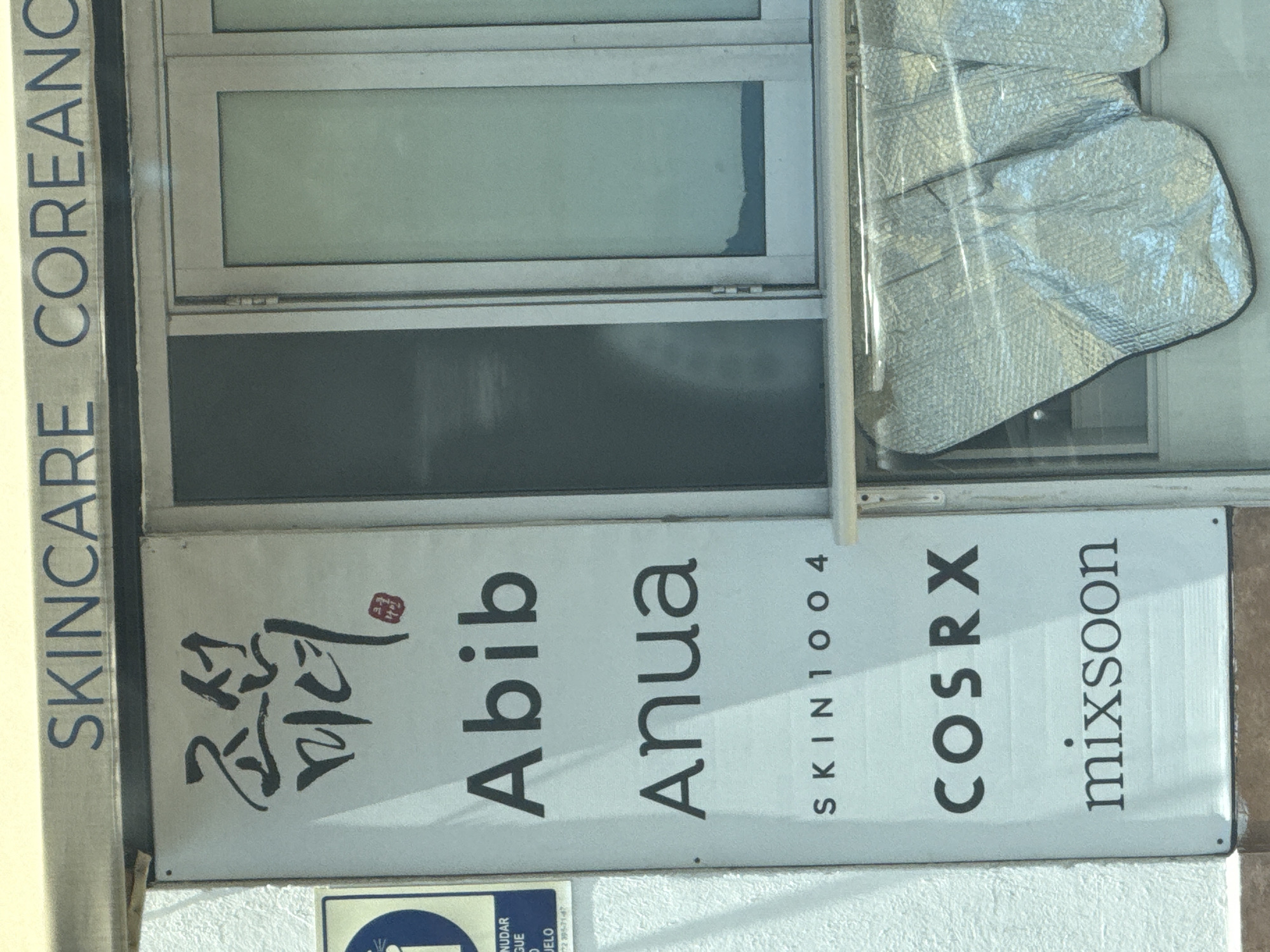 The image shows a storefront sign for a Korean skincare business. The sign lists brands such as "Abib," "Anua," "COSRX," and "Mixsoon." The phrase "SKINCARE COREANO" (Korean Skincare) is vertically oriented along the left edge of the image. The sign is located in the city of Puebla, Mexico. There is a reflective sunshade visible through the store window.