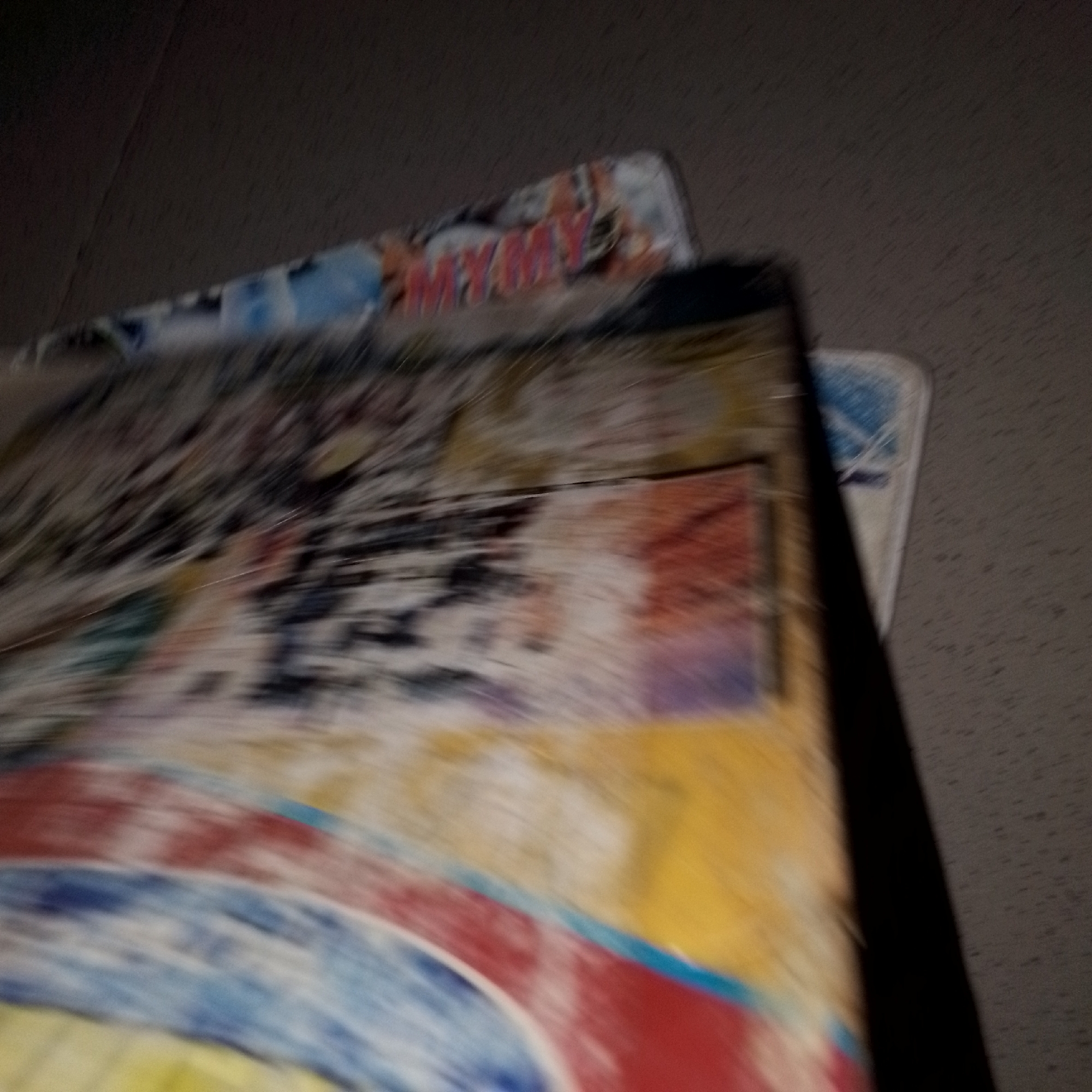The image displays a close-up, blurred view of what appears to be stacked notebooks or folders with colorful covers. One cover has the word "MY" repeated in red lettering, and the overall design features abstract patterns in blue, red, yellow, and white. The items are angled, with the lower one appearing to be a binder or thick book. The background is dark and textured, suggesting a floor or wall. There are no people or specific landmarks visible that would indicate a location in Jalingo, Nigeria, or any other city. The image is of poor quality due to motion blur and low light, making precise identification of details difficult.
