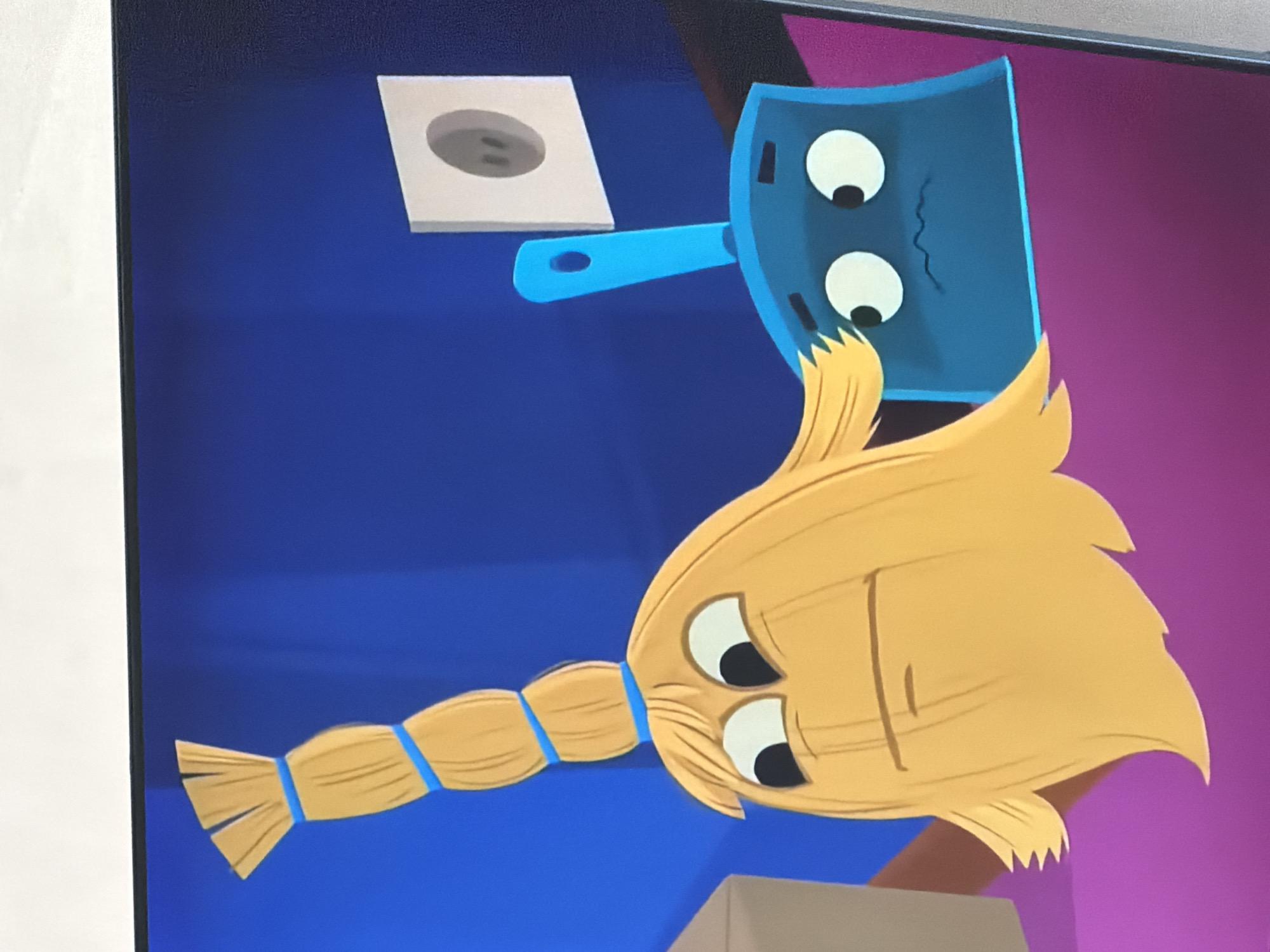 The image is a close-up of an animated scene featuring two anthropomorphic cleaning tools against a blue background. On the left is a yellow broom with a segmented handle tied with blue bands and a head of bristly yellow strands that form a face with wide, surprised eyes and a straight mouth. Next to it on the right is a blue dustpan with a similar cartoon face, featuring large, round white eyes with black pupils and a small, wavy, distressed mouth. A light gray electrical outlet with two dark gray holes is mounted on the blue wall to the right of the broom. The lower portion of the scene shows a purple surface, and to the far left, a corner of a brown cardboard box is visible. There is no visible connection to the city of Krasnyi Krym, Russia.