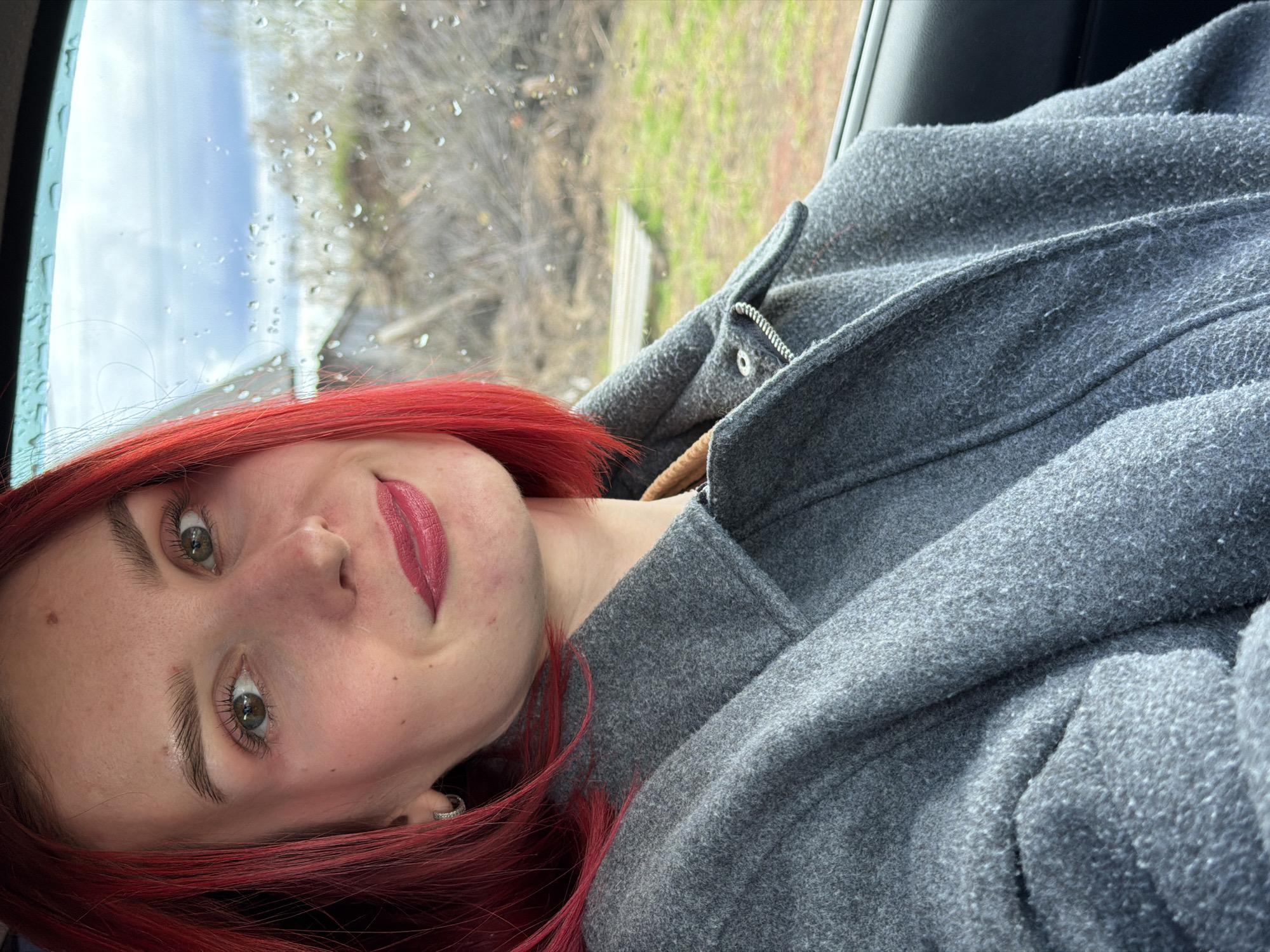 A selfie shows a woman with red hair wearing a gray coat in a car with rain-streaked windows. Outside the window, trees and buildings are visible, suggesting an urban setting, likely in Chaikovsky, Russia. The woman is looking at the camera with a slight smile. The lighting is natural, indicating daytime.