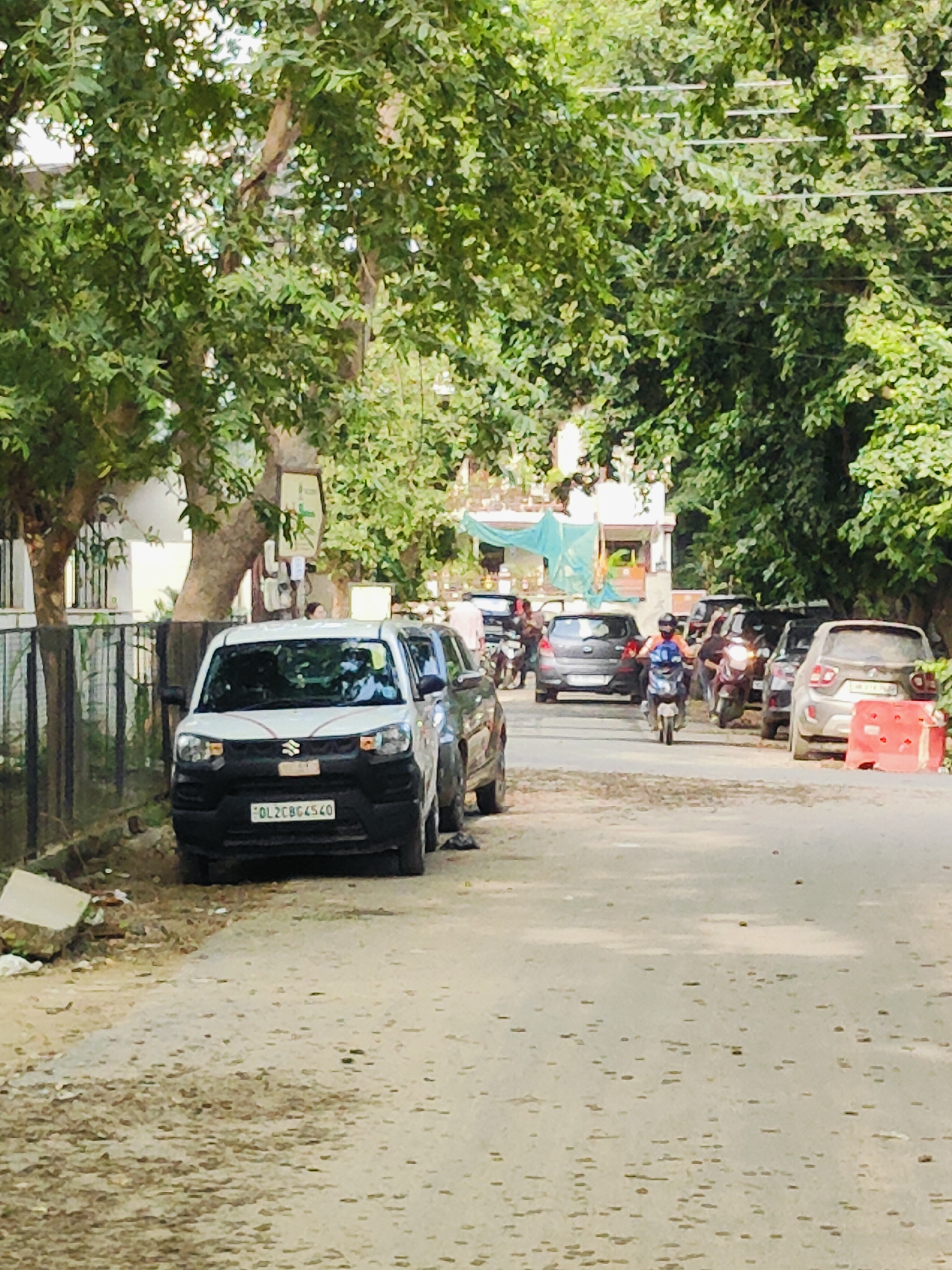 A white Maruti Suzuki car is parked on a dusty road lined with trees in Gurgaon, India. The car, license plate DL2CBG4540, is facing the camera. Behind it, several other vehicles, including a dark hatchback and a silver SUV, are also parked or in motion on the road. A person on a scooter is visible in the middle of the road, heading away from the camera. The environment appears to be a residential street with buildings and lush green trees providing shade. Red and white traffic barriers are present on the right side of the road.