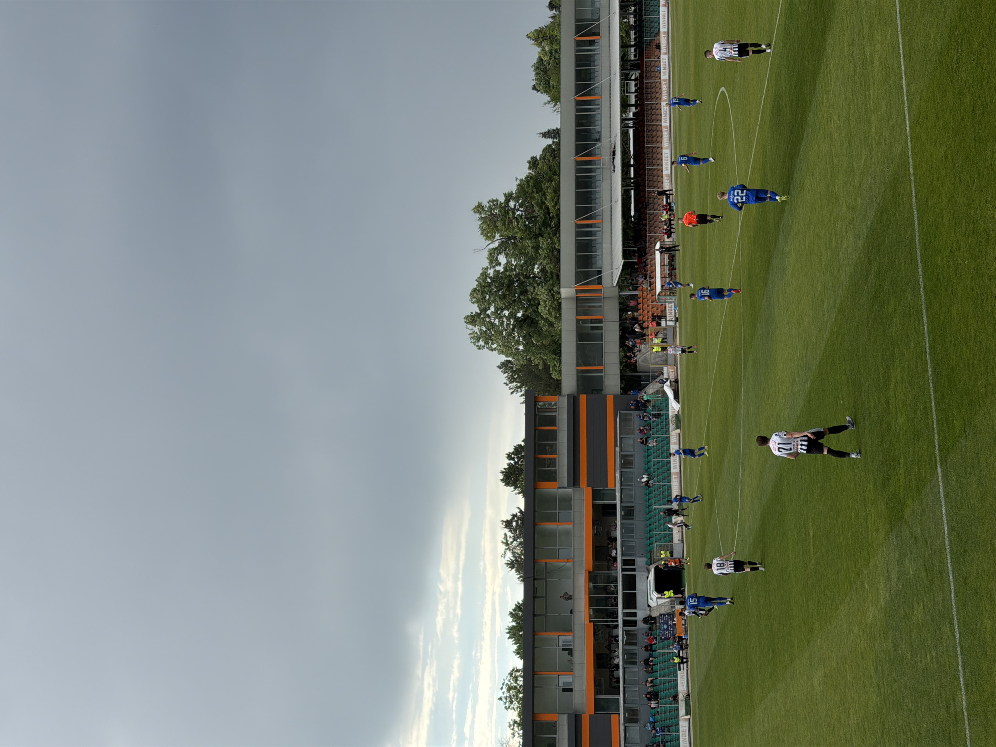 The image shows a soccer game in progress. The game is being played on a green field, with players in blue and white-and-black uniforms. Spectators can be seen in the stands of a multi-story building with orange and grey accents. The sky is overcast with both cloudy and clear areas. This likely depicts a game at the Lovech Stadium in Lovech, Bulgaria.