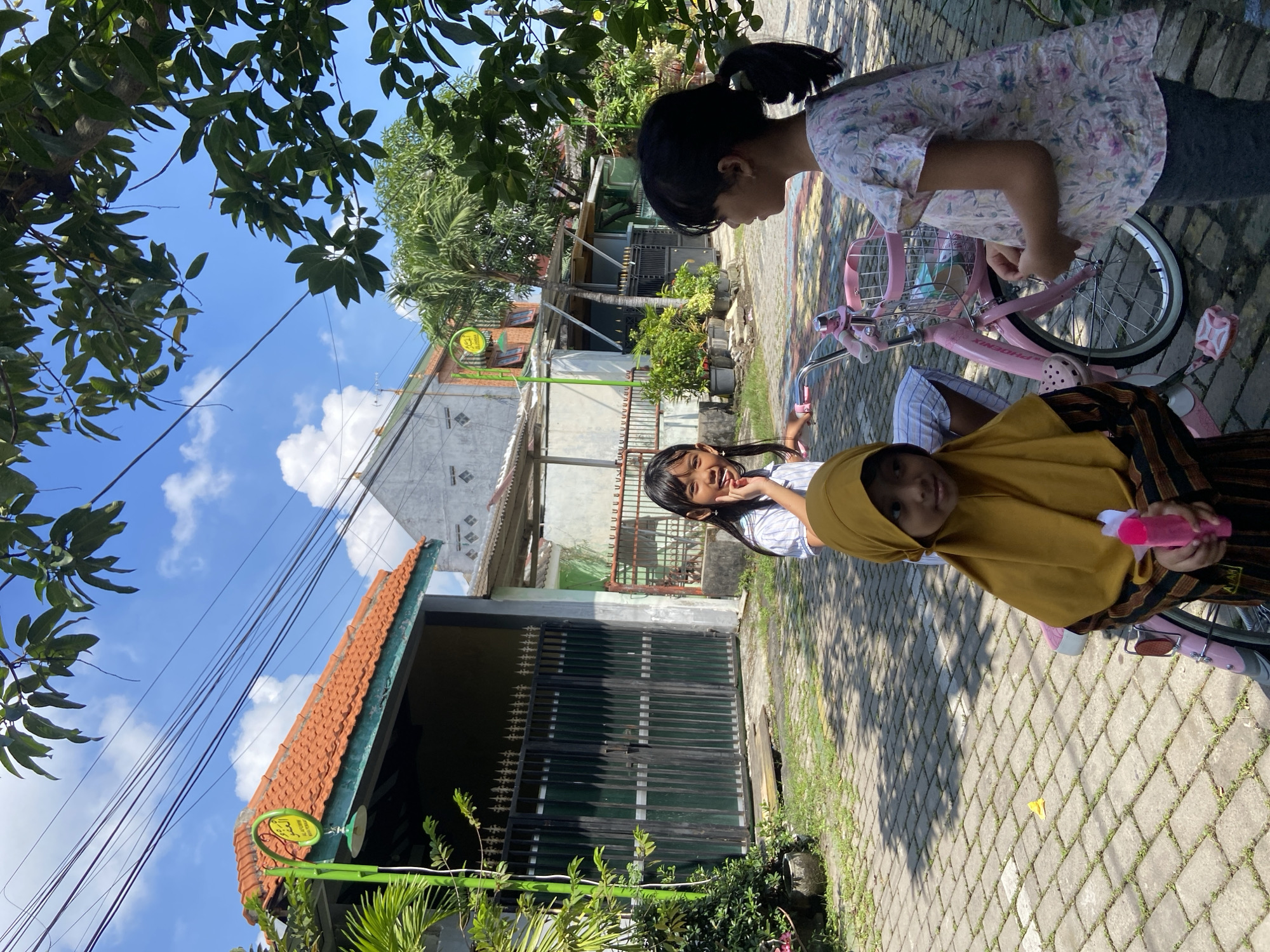 Three young girls are on a brick-paved path in Gelam, Indonesia. One girl, wearing a yellow hijab and striped shirt, sits on a pink bicycle, holding a pink bottle. Another girl stands beside her, smiling and looking at the camera. A third girl with a ponytail leans over the handlebars of the bicycle. Behind them are houses and fences, with greenery and trees visible. The sky is blue with scattered clouds.