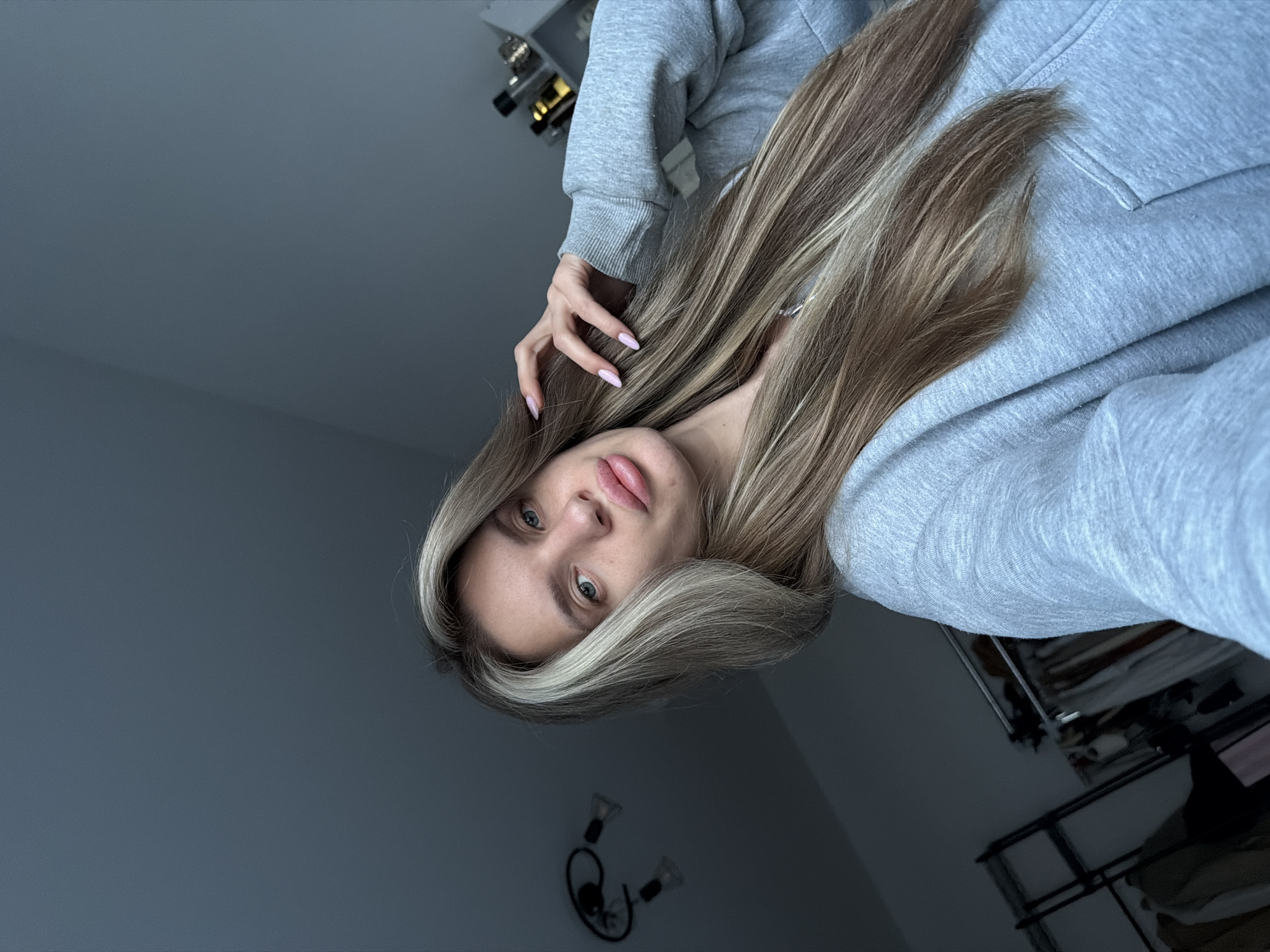 A young woman is centered in the image, wearing a gray hooded sweatshirt. She is looking at the camera from a tilted perspective. Her long, highlighted blonde hair partially obscures her face and her right hand is near her hair. The background is a muted gray tone, with a shelf containing various items visible in the top left corner and a rack with what appears to be clothes in the lower right. A small, minimalist lamp fixture is seen on the wall in the lower portion of the image. There is no information in the image to determine if this is Berlin, Germany.
