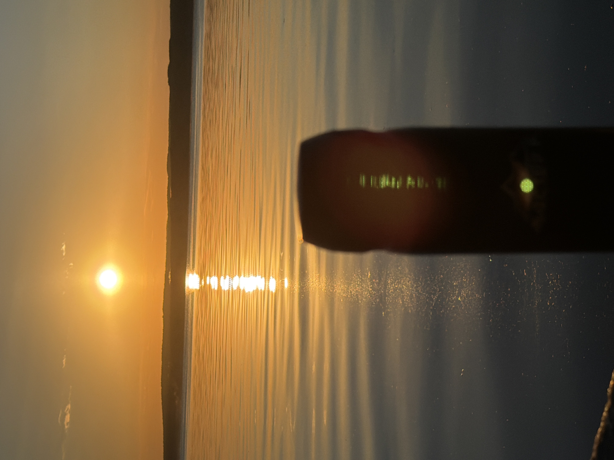A silhouetted object stands in the foreground, out of focus, against a tranquil body of water reflecting the warm, golden hues of a sunset. The sun, a bright orb, is positioned high in the sky, casting a brilliant reflection on the rippling water that stretches towards a dark, silhouetted horizon line. The sky above transitions from a deep orange near the horizon to a paler shade higher up, with a few wispy clouds. This scene captures the serene beauty of a sunset over the Kama River in Chaikovsky, Russia.