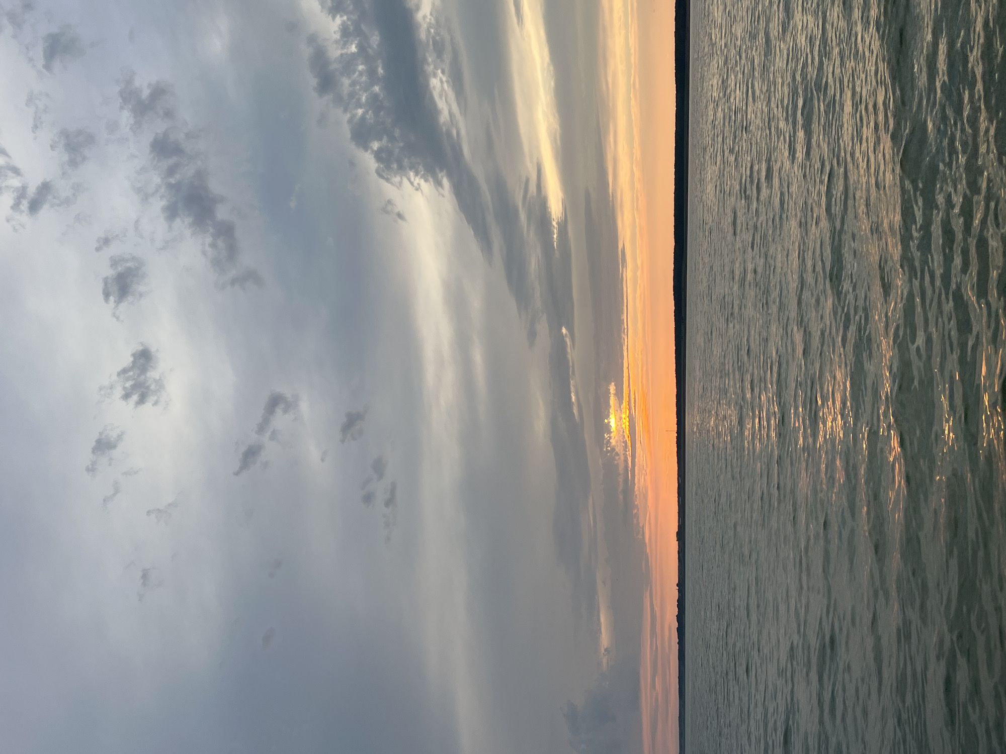 The image depicts a serene body of water under a cloudy sky at sunset. The horizon is marked by a distant shoreline with dark trees and a few structures, possibly a small town or settlement. The sky transitions from muted grays and blues at the top to vibrant oranges and yellows near the horizon, where the sun is partially visible, casting a warm glow on the clouds and reflecting on the water's surface. The water itself appears calm, with gentle ripples creating a shimmering effect from the fading sunlight. The scene evokes a sense of tranquility and the natural beauty of a sunset over a body of water. The location is not specified as Borovoe, Russia; however, the overall landscape suggests a natural setting, likely a lake or a large river, conducive to such sunset views. There are no people or specific identifiable objects in the foreground or middle ground, emphasizing the natural environment.