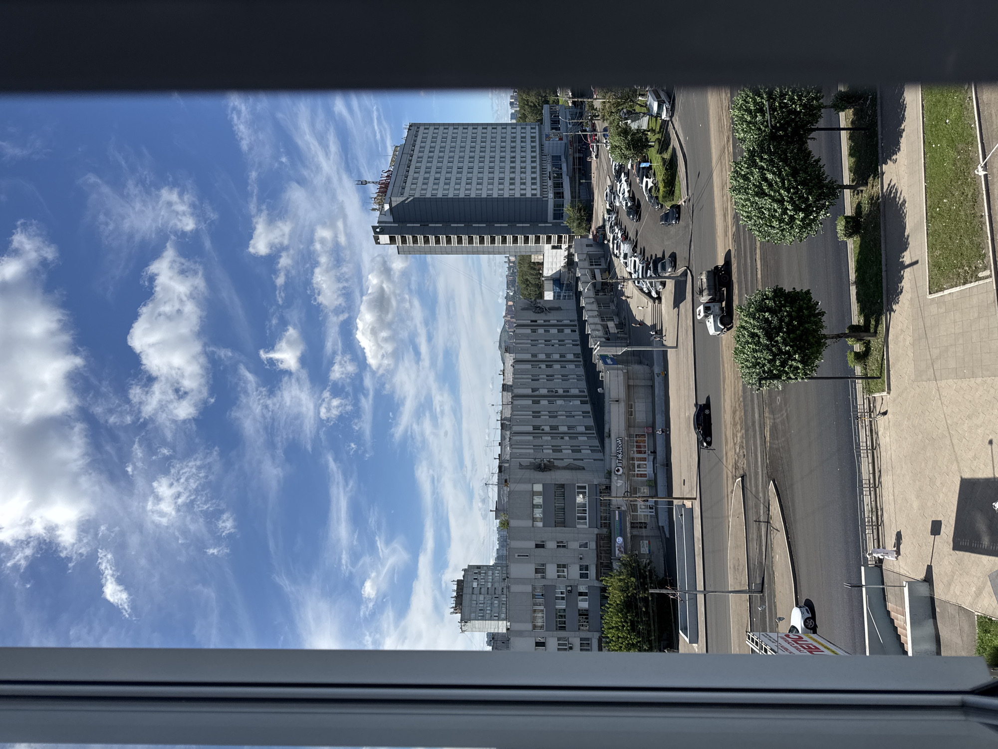 This is a high-angle, eye-level, full shot of a city street and buildings on a sunny day with scattered clouds in Krasnoyarsk, Russia. In the foreground, there's a white framed window on the left and a dark framed window on the right. The middle ground shows a street with several cars parked on the right side. There are also trees lining the street and a white truck is driving on the road. In the background, there are several apartment buildings and a tall hotel. The sky is blue with white clouds.