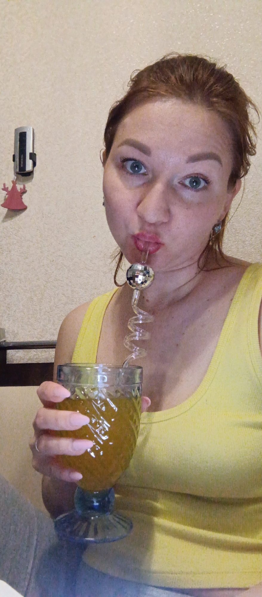 A woman with red hair pulled back is posing with a glass filled with a yellow-brown liquid and a coiled glass straw with a disco ball on the end, she has her lips pursed around the straw. She is wearing a yellow tank top. A silver device and a red holiday decoration are mounted on the wall behind her. The location is unknown.