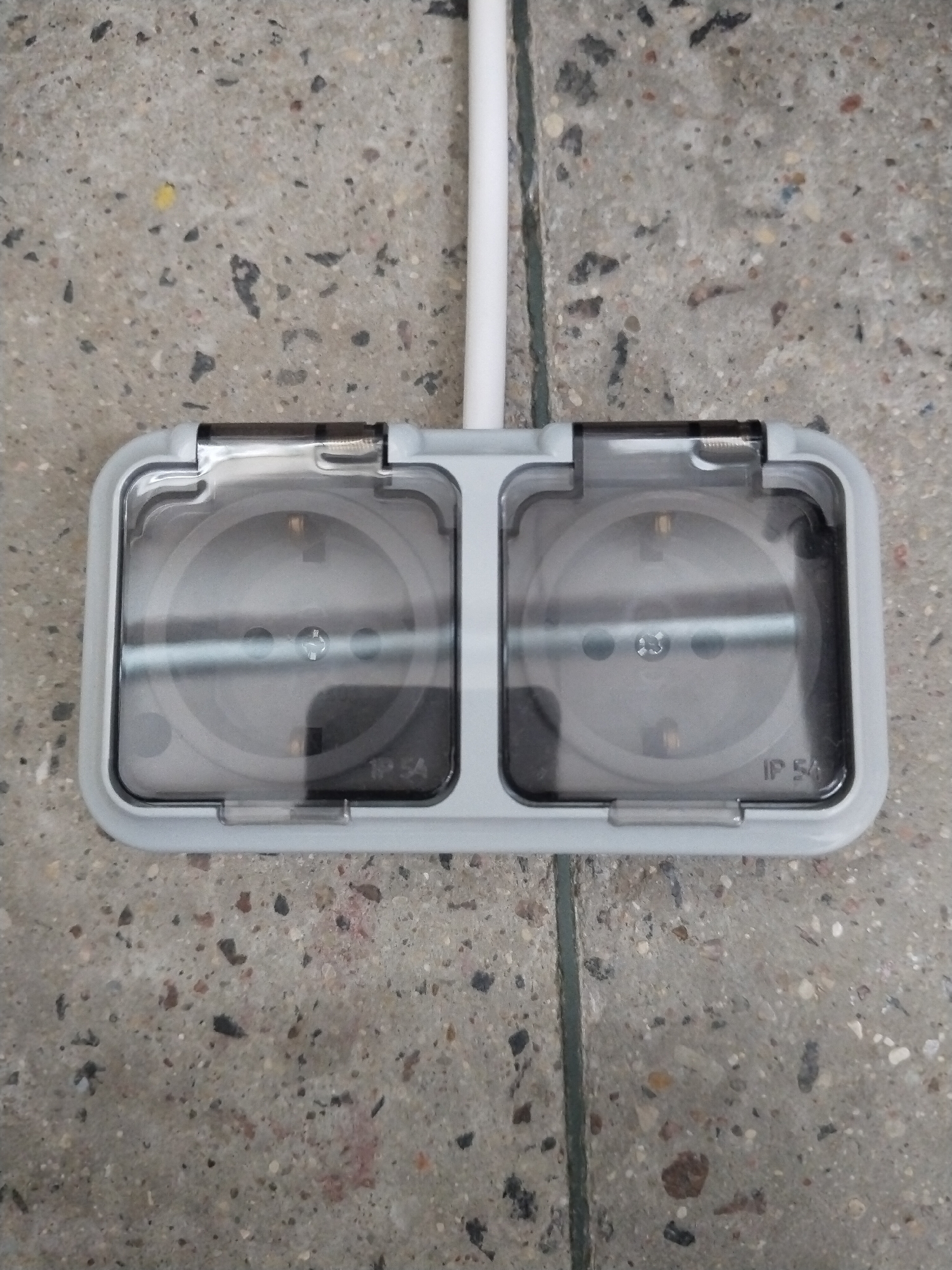 An outdoor, double electrical socket is mounted on a speckled concrete surface. A white conduit extends vertically upwards from the top of the socket. Each socket is protected by a clear, hinged plastic cover. The socket housings are light grey. The right-hand socket has "IP 54" printed on its cover. The concrete appears to be part of a sidewalk or paved area, with a visible seam running vertically down the center.