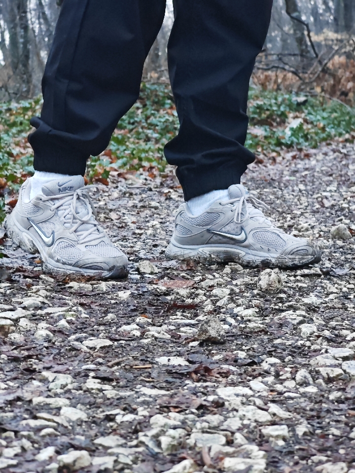 A person is depicted from the lower legs downward, standing on a natural, unpaved path. The individual wears black athletic pants with elasticized cuffs, white socks, and a pair of light grey Nike athletic shoes. The shoes, with visible white laces, show accumulation of mud and dirt on their soles and sides. The path beneath consists of irregular light-colored rocks, dark soil, and scattered damp, brown leaves. Some green ground cover is visible along the edges of the path and in the blurred background, which shows indistinct trees, indicating a wooded environment. This scene is located on a forest trail within the Shumen Plateau Nature Park, bordering the city of Shumen, Bulgaria.