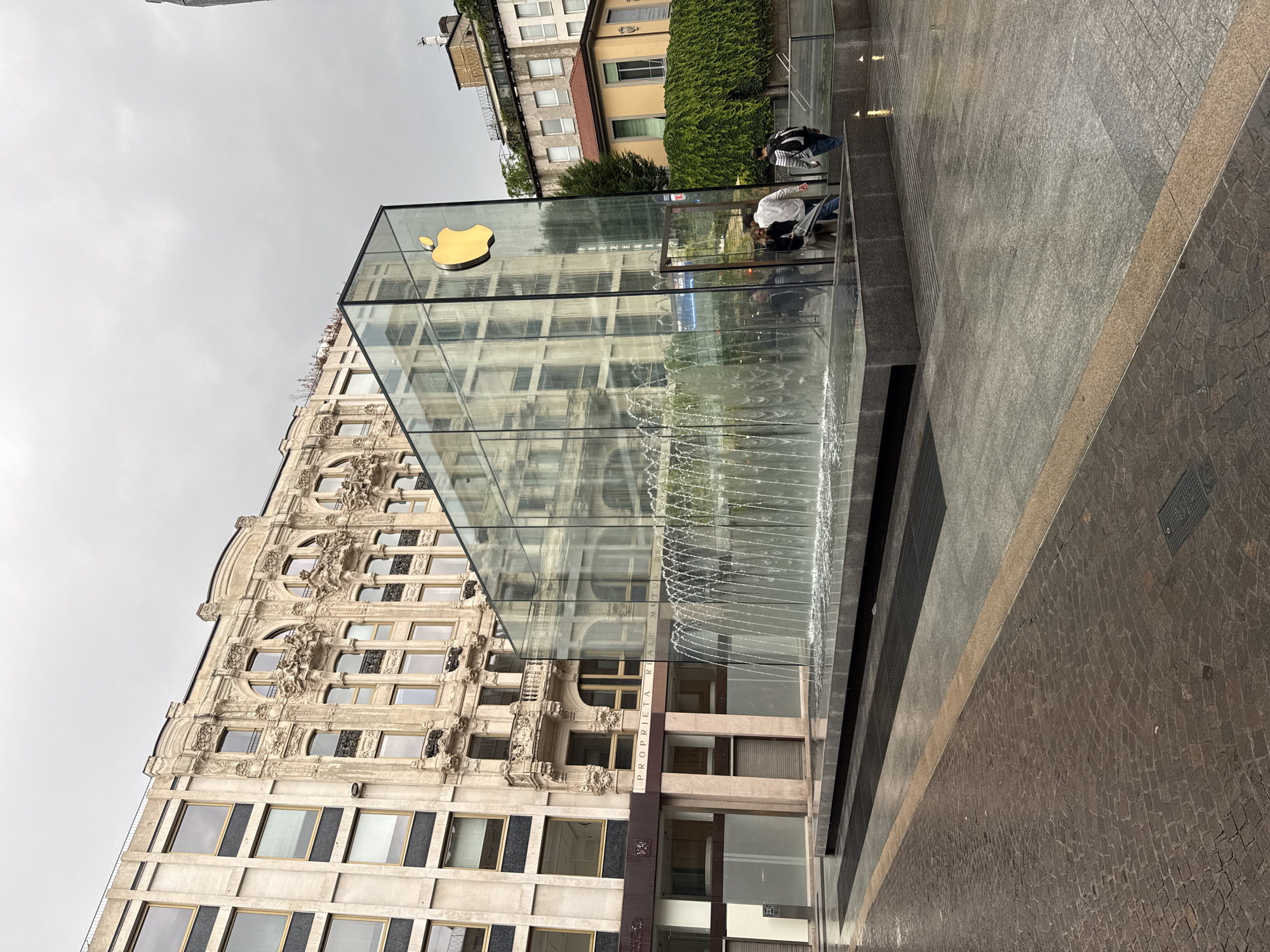 This image captures the distinctive Apple Piazza Liberty store in Milan, Italy, under an overcast sky during daytime. The central feature is the store's unique entrance: a large, transparent glass cube adorned with a prominent golden Apple logo on one side. A striking water feature forms a continuous cascading fountain down the front side of the glass cube, creating a watery barrier and an immersive entry experience.
To the left of the glass structure stands an elegant, classical-style building characterized by its light-colored, ornate facade. This building boasts multiple windows and intricate architectural details, including decorative carvings, reflecting a historical European design. Further back and to the right, another, more modern building with simpler lines and some lush green foliage is partially visible.
Several individuals are present in the scene. Two people are standing near the glass entrance of the Apple Store, appearing to observe or approach the entrance, dressed in casual attire. The ground in the foreground is a textured paved area, likely a pedestrian square, with a pattern of grey and brown cobblestones or tiles.
A narrow brown band on the building directly to the left of the glass cube, beneath some windows, displays the text "PROPRIETA REALE MILANO". The overcast sky lends a soft, diffused light to the scene, highlighting the contrast between the modern glass and water structure and the surrounding historical architecture in a vibrant urban setting.