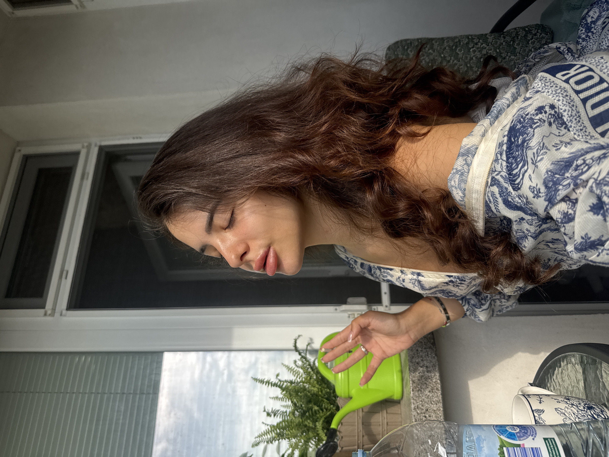 A young woman with long, wavy, dark brown hair is captured in a medium shot, seated on a balcony. She is wearing a white top with blue intricate patterns and a zipper, partially unzipped. Her eyes are closed, and her face is tilted upwards towards the sun, with her lips slightly parted. Her right hand is raised near her face, with fingers slightly curled, revealing a ring on her ring finger. She is wearing a bracelet on her left wrist.
In the background, to the left of the woman, a bright green watering can sits on a windowsill. Behind the watering can, a potted fern is visible. To the far left, a partially visible plastic water bottle with a blue label stands upright. A white mug with blue floral patterns is on a glass-top table in the foreground. The scene appears to be outdoors on a balcony, with a window and its blinds to the left, and a wall to the right. The lighting suggests it is daytime, with sunlight illuminating the woman's face. The overall setting does not provide any specific visual cues that would pinpoint the location to Plovdiv, Bulgaria.