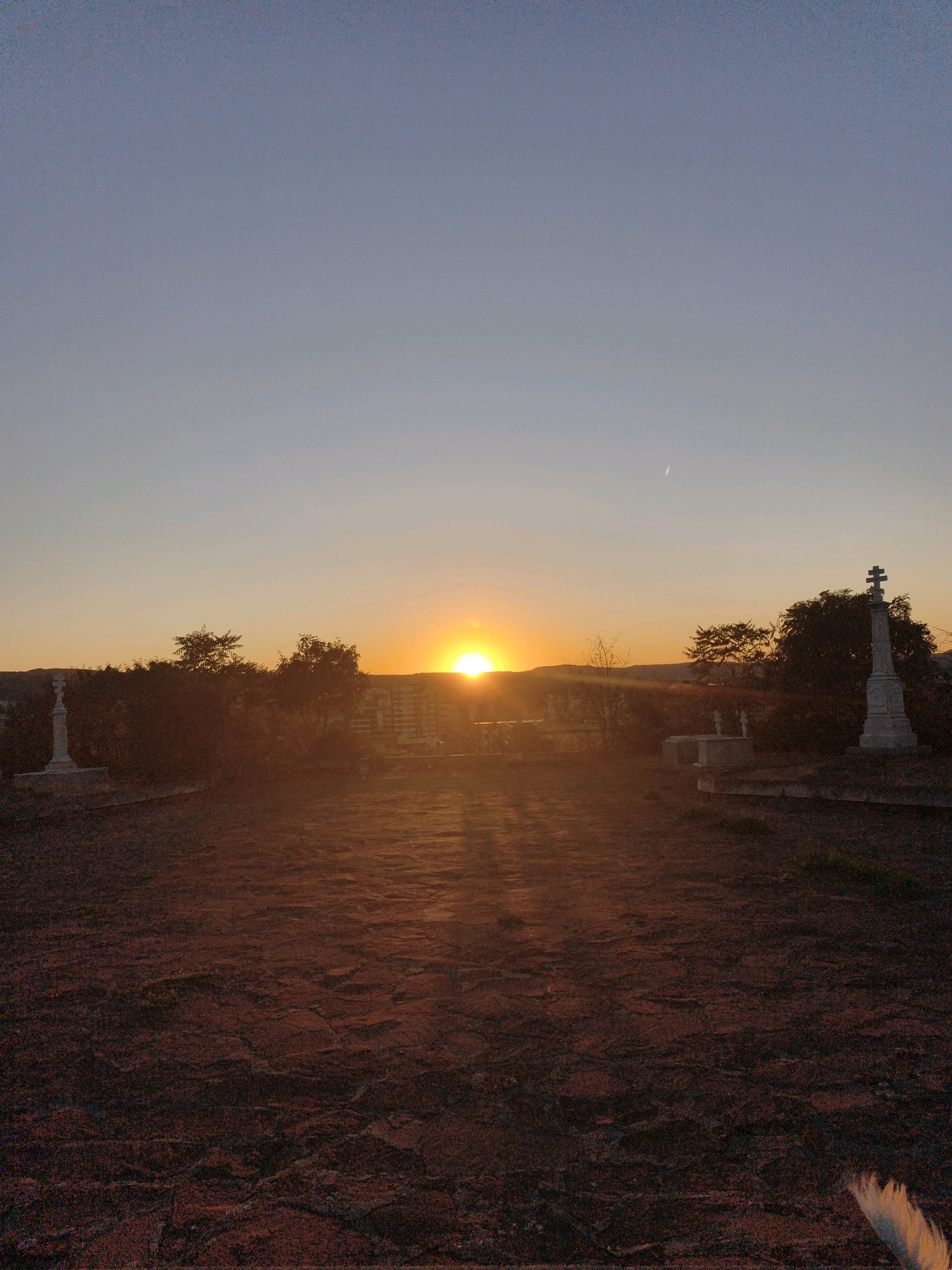 The image depicts a sunset over the city of Lovech, Bulgaria, viewed from a cemetery. In the foreground, a stone-paved path, possibly weathered, is illuminated by the setting sun, casting long shadows. The path leads towards a line of trees and a cityscape in the distance. Several gravestones and monuments are visible, including a prominent white cross-shaped monument on the right and another stone structure on the left. The sky transitions from a clear blue at the top to a warm orange and yellow hue near the horizon where the sun is setting. The overall atmosphere is serene and somber, characteristic of a cemetery at dusk.