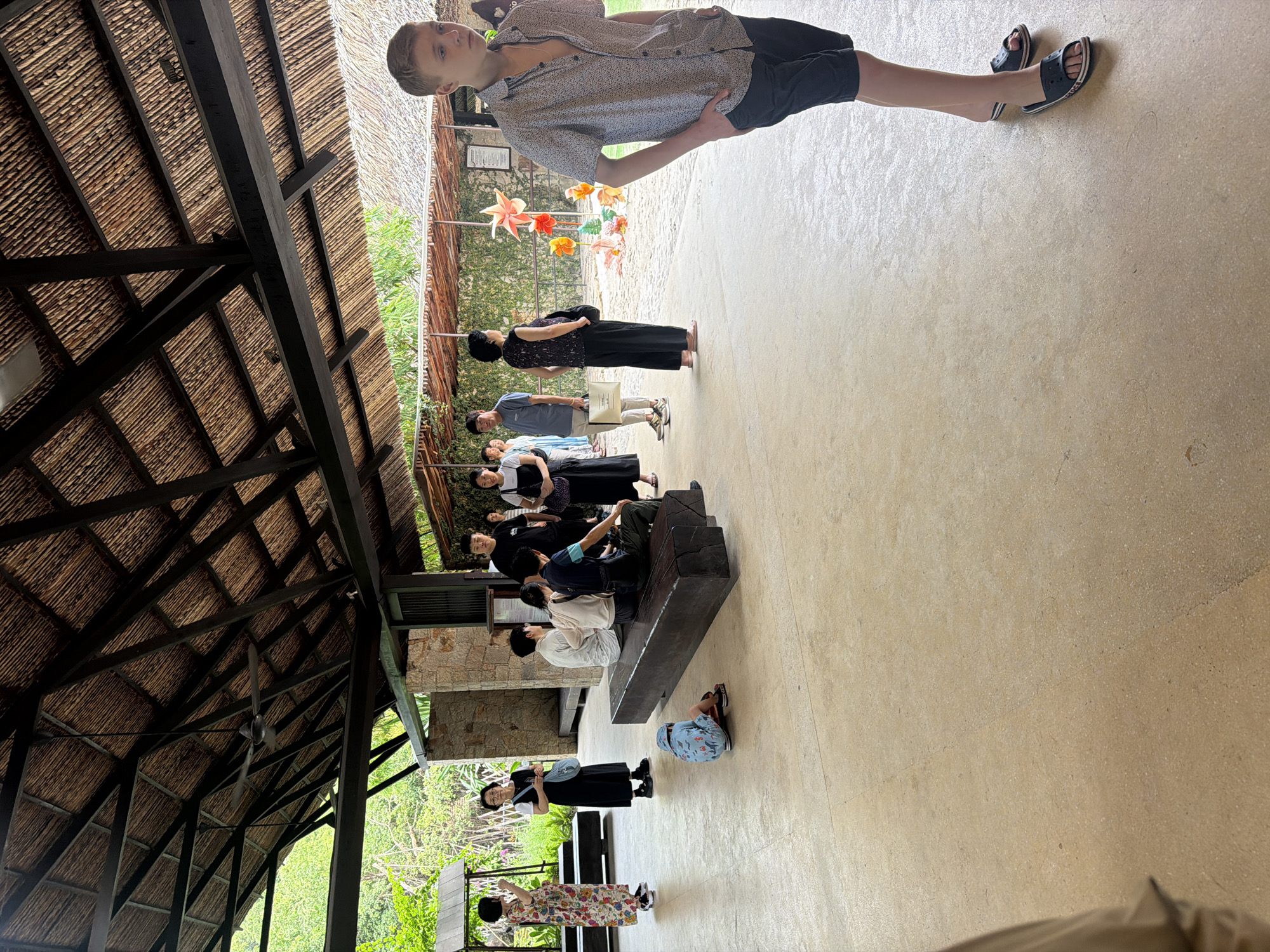 The image depicts an outdoor covered seating area, likely part of a tourist attraction or resort, in Nha Trang, Vietnam. The architecture features a thatched roof supported by dark wooden beams, creating a rustic and tropical ambiance.
In the foreground, a young Caucasian male with blond hair, wearing a patterned short-sleeved shirt and black shorts, stands facing left with his hands in his pockets, looking out of frame. He is wearing black slides with white trim.
To the left, a group of people are gathered. A young child in a blue patterned outfit is sitting on the ground, facing away from the camera. Further left, several adults are standing or sitting. Two women, one in a long floral dress and another in a black dress, are standing and facing right. Behind them, a group of people, including men and women, are standing and appear to be looking at something off-camera. One man is wearing a black t-shirt and dark pants, while another is in a light-colored t-shirt and beige pants, holding a white paper bag. A woman in a black sleeveless top and black pants is also present.
In the mid-ground, a long wooden bench is occupied by several individuals, some sitting and some standing. A few people appear to be interacting or observing something in the background.
The background reveals a lush green environment with trees and foliage, suggesting a tropical setting. Decorative, colorful flower-like structures are visible, possibly part of an exhibition or art installation. The ground surface appears to be a light-colored, smooth concrete or stone material. A ceiling fan is visible attached to the wooden beams of the roof. The overall scene suggests a relaxed, possibly tourist-oriented environment.