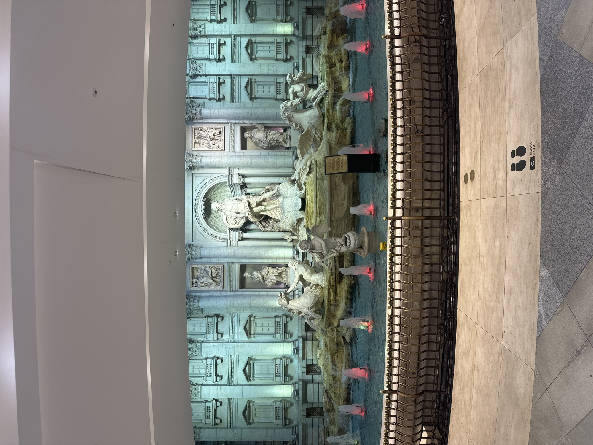 The image captures a replica of the Trevi Fountain, situated in the city of Seoul, South Korea. The fountain, a detailed imitation of the original, features classical statues and architectural elements, with water illuminated by red lights. A metal barrier separates the fountain from a tiled pedestrian area. "Photo Zone" markers are on the ground, indicating designated photo spots. The ceiling architecture of an indoor space is visible above the fountain. No people are in the visible portion of the image.