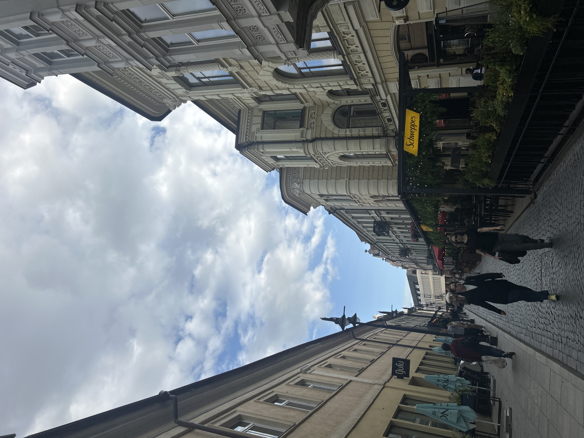 The image shows a view down a cobblestone street in Vilnius, Lithuania. The street is lined with tall, ornate buildings. On the left side, a yellow building with the "Gulu" logo is visible, with blue umbrellas in front of it. On the right side, a building with a Schweppes sign is prominent, with outdoor seating for a cafe. Several people are walking on the street, with two women in the foreground walking towards the viewer. The sky is partly cloudy.