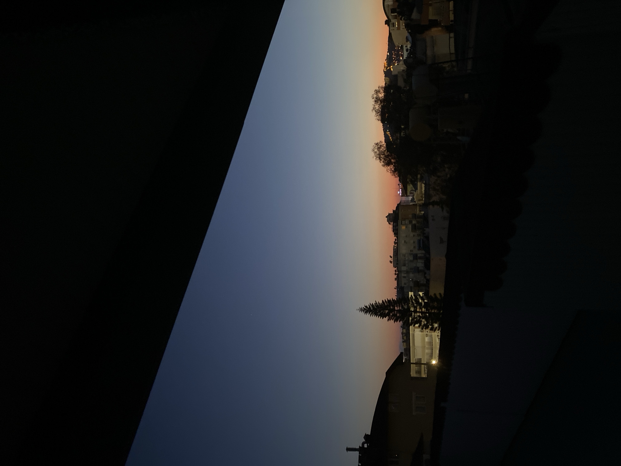 The image captures a serene twilight view of a cityscape, likely Alanya, Turkey, from an elevated perspective. The sky dominates the upper portion of the frame, displaying a gradient of colors characteristic of dusk. It transitions from a deep indigo and dark blue at the top to softer hues of orange and pink along the horizon line.
Silhouetted against the fading light, numerous buildings, houses, and other urban structures form a dense cityscape across the lower half of the image. Scattered artificial lights within the buildings begin to illuminate, suggesting the transition into night. Interspersed among the structures are the dark silhouettes of trees and other vegetation, including a prominent evergreen-like tree in the mid-ground.
A large, dark, diagonal area occupies the left side of the frame, partially obscuring the view. This element appears to be an interior feature, possibly a window frame or part of a building from where the photo was taken. The weather appears clear, with no clouds visible. The overall atmosphere is peaceful and calm, highlighting the quiet beauty of the city as day turns to night. There is no visible text or explicit human activity in the scene.