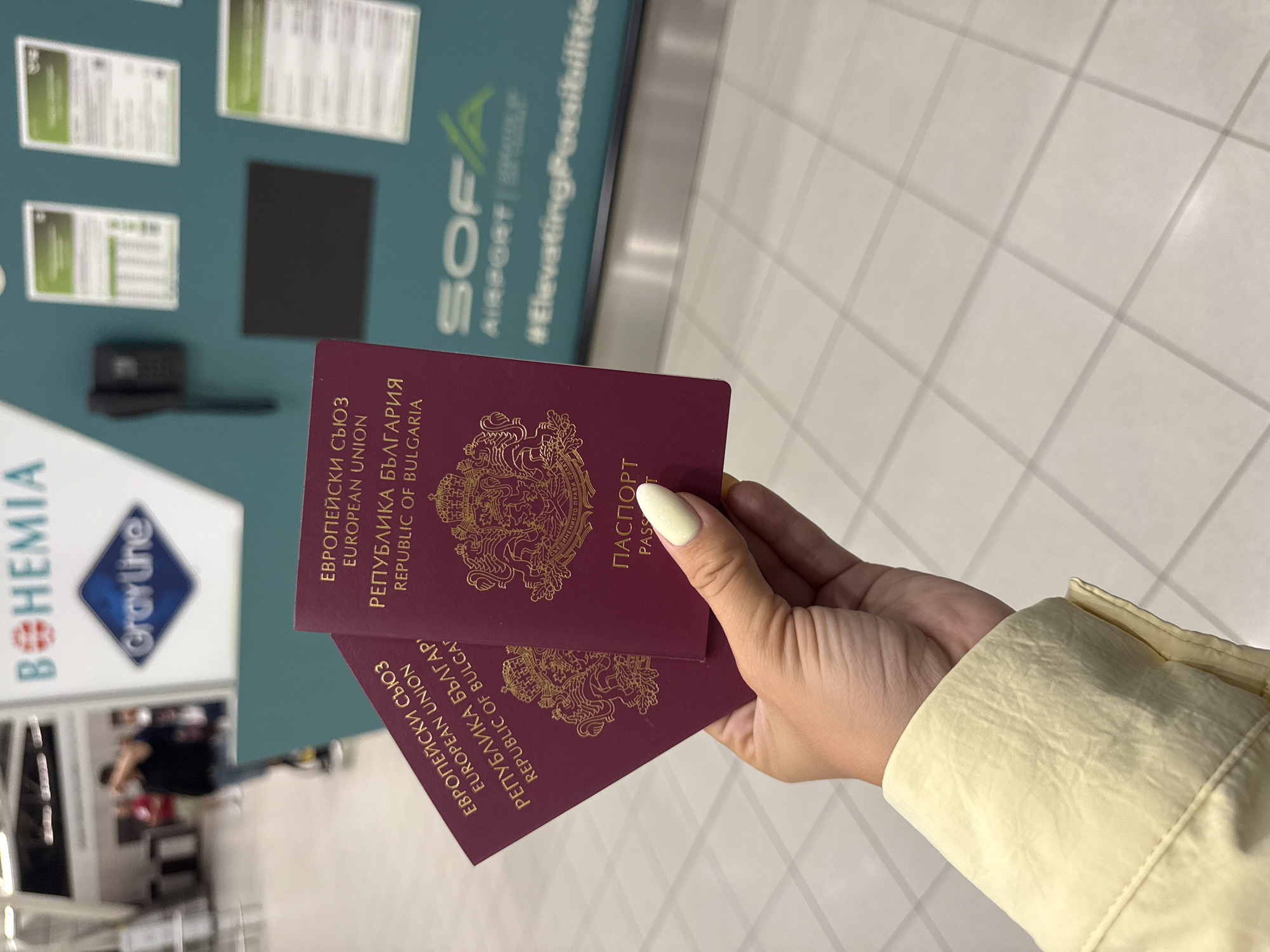 A hand with light-colored fingernail polish holds two burgundy Bulgarian passports at the SOF Airport in Sofia, Bulgaria. The passports are open to pages displaying the Bulgarian coat of arms and text in Bulgarian and English. In the background, a sign reads "SOF AIRPORT" and "#ElevatingPossibilities". A person can be seen in the blurred background.
