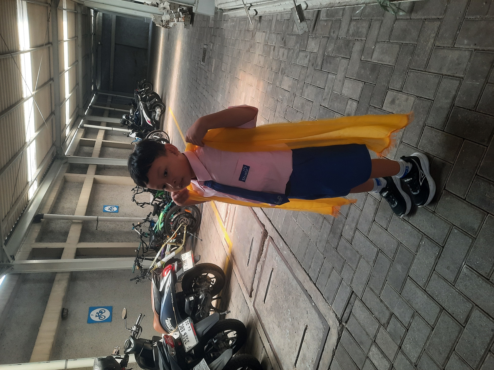 A young boy wearing a pink uniform shirt, blue shorts, a dark blue tie, and black sneakers stands in a covered outdoor area. He has a yellow cape draped over his shoulders, with one hand holding it up near his neck. The boy is positioned on a paved walkway made of interlocking gray bricks, with a distinct herringbone pattern. Behind him, several motorcycles and bicycles are parked in rows, suggesting a parking area. The walls of the surrounding structure are made of textured concrete. Two blue signs with white bicycle symbols are visible on the wall to the left. The ceiling is a corrugated metal structure with translucent panels allowing natural light to filter through. The scene appears to be in an urban environment, likely a public space or a facility in North Jakarta, Indonesia, possibly a school or a multi-purpose complex. The lighting suggests it is daytime.