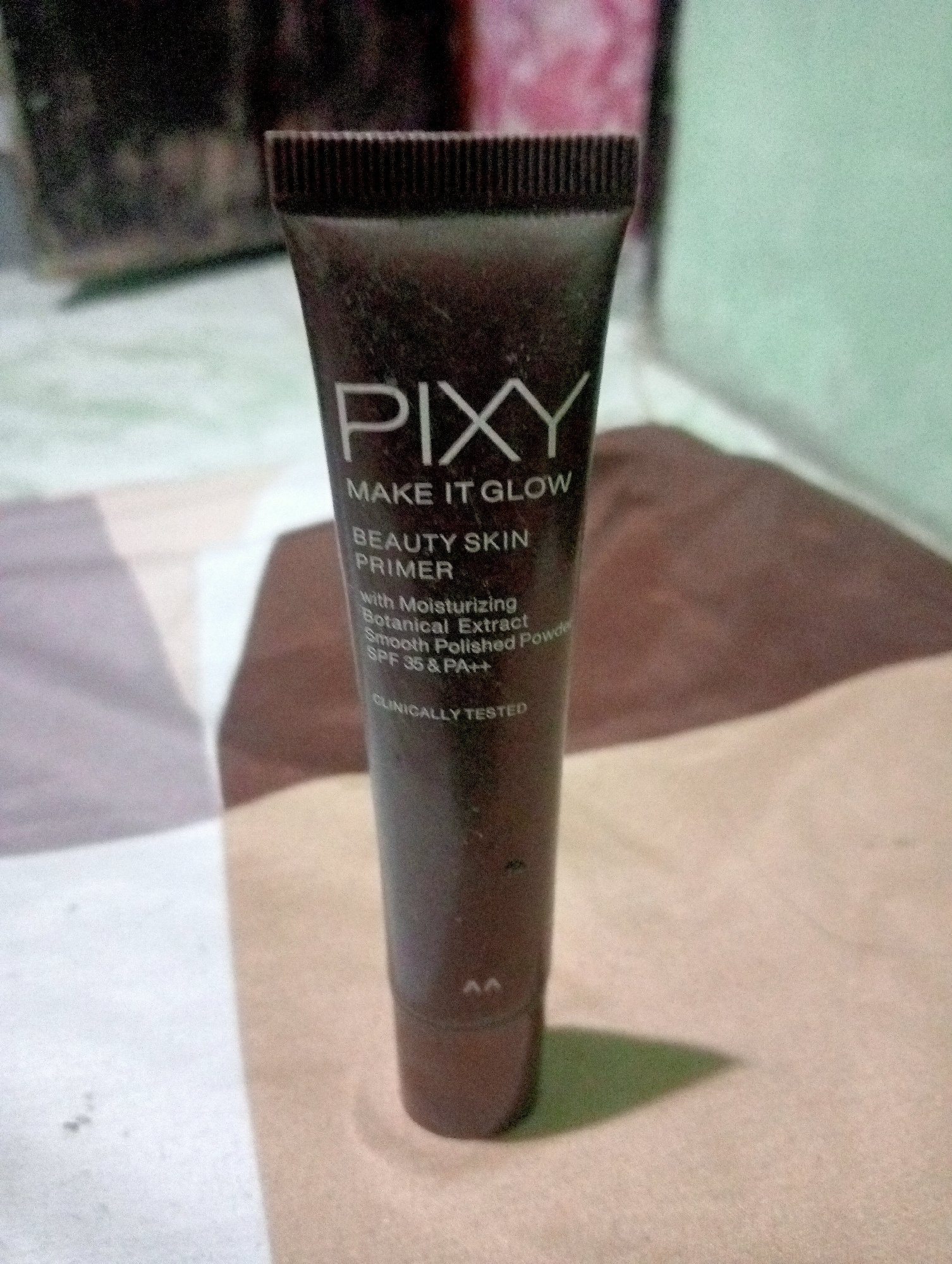 A tube of PIXY MAKE IT GLOW BEAUTY SKIN PRIMER is positioned vertically in the center of the frame, resting on a textured surface that appears to be a bedspread or mat. The tube is dark brown with white text. The text on the tube reads "PIXY", "MAKE IT GLOW", "BEAUTY SKIN PRIMER", "with Moisturizing Botanical Extract", "Smooth Polished Powder", "SPF 35 & PA++", and "CLINICALLY TESTED". The cap of the tube is ribbed and dark brown. A shadow of the tube is cast to the right on the surface. The background is out of focus and displays colors that suggest an indoor setting, possibly a room. There is no indication of the city of Kota Negara Timur, Indonesia in the image or its context.