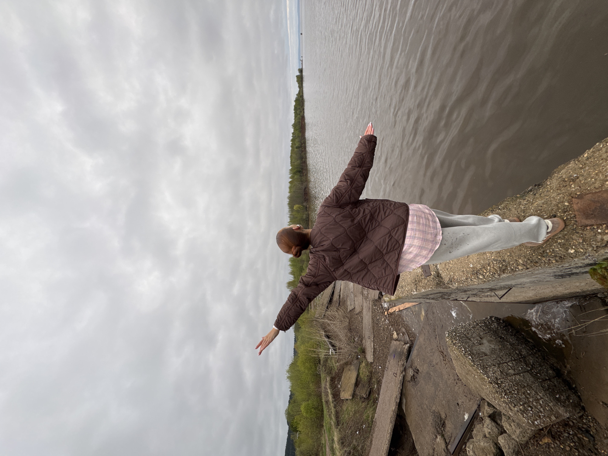 A woman is standing on a concrete structure near a body of water. She is wearing a brown quilted jacket, a light pink and white plaid shirt, and gray sweatpants. Her arms are outstretched to the sides. The water is calm and gray, and across the water, there is a line of trees. The sky is overcast with gray clouds. Below the concrete structure, there is vegetation. The location is Chastyye, Russia.