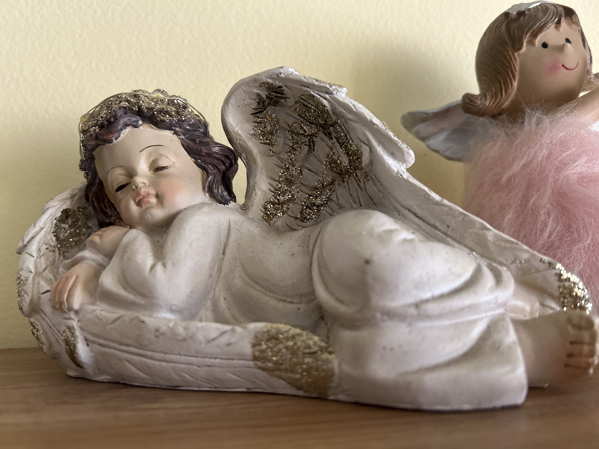 Two angel figurines are on a wooden surface, likely a shelf. The foreground figure is a sleeping angel with brown hair and gold glitter accents on the hair and wings. The angel is in a reclining position with eyes closed, wearing a white robe-like garment, and resting on a feather-shaped base. The background figure is a standing angel with brown hair, a pink fluffy dress, and small wings. The background is a light yellow wall. The location is unknown.