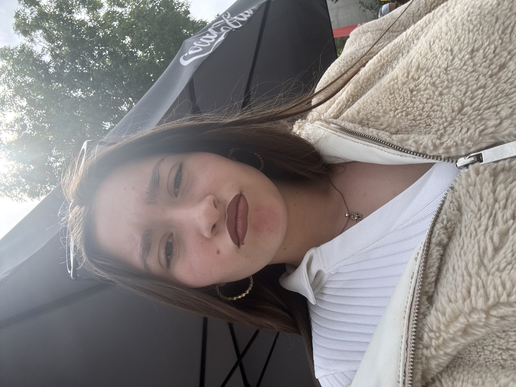A young woman is posing for a selfie. She has long brown hair, dark eyebrows, and is wearing lipstick. Her clothing consists of a white shirt and a fuzzy tan jacket. She has hoop earrings and a necklace. Above her and to the left, there is a black Coca-Cola-branded umbrella. A tree is visible in the background. There is insufficient information to determine the exact location in Sofia, Bulgaria.