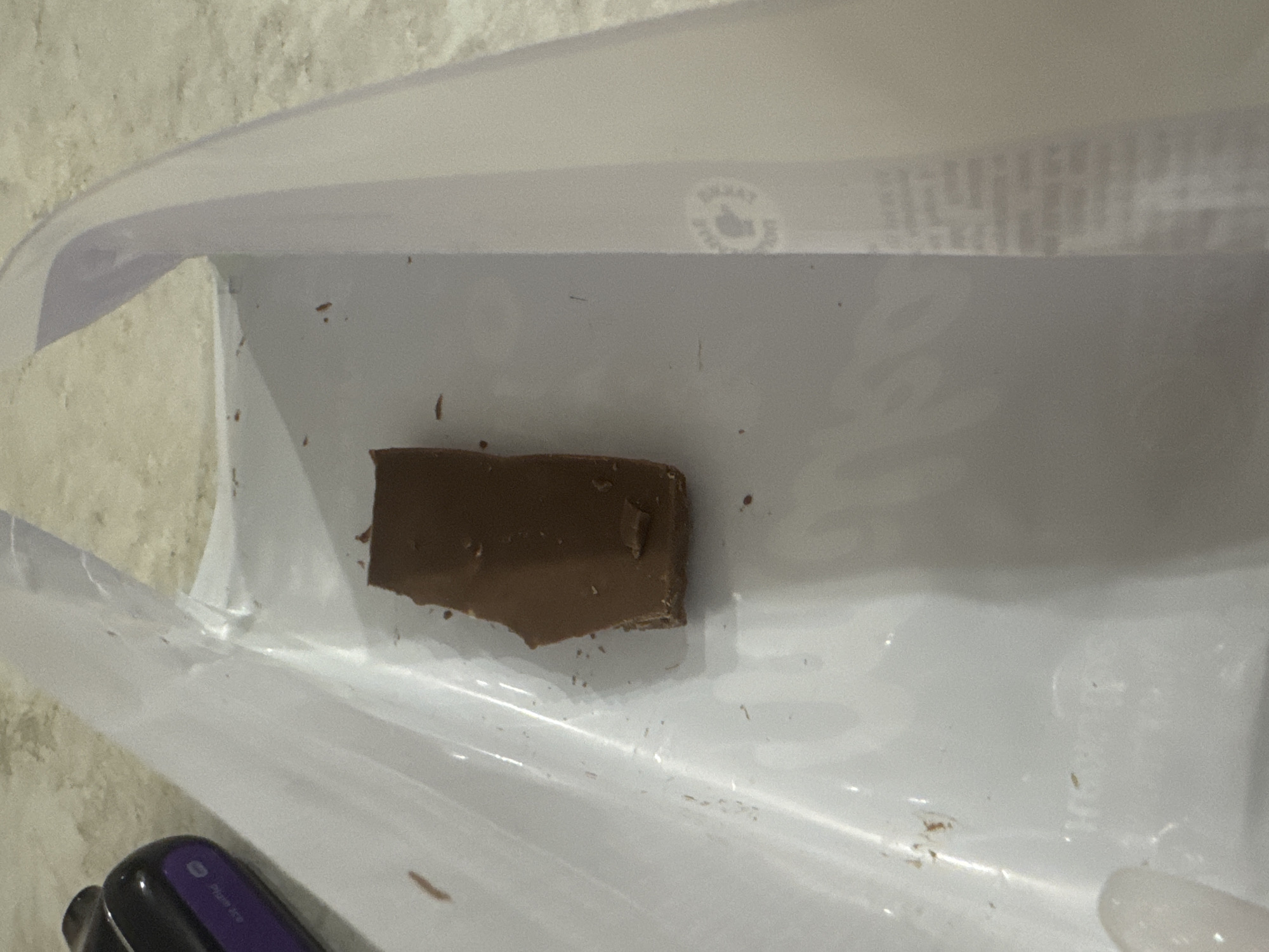 The image shows a close-up, overhead view of a rectangular piece of dark chocolate resting inside a clear plastic container. The chocolate is broken and irregular in shape, with a bite taken out of one end. The container is transparent and appears to be a food storage box, with some smudges and minor debris scattered on its surface. The background suggests a countertop or table surface. The scene is indoors and there is no indication of the location being in Saratov, Russia. The image is focused on the chocolate piece and the immediate surroundings within the container.