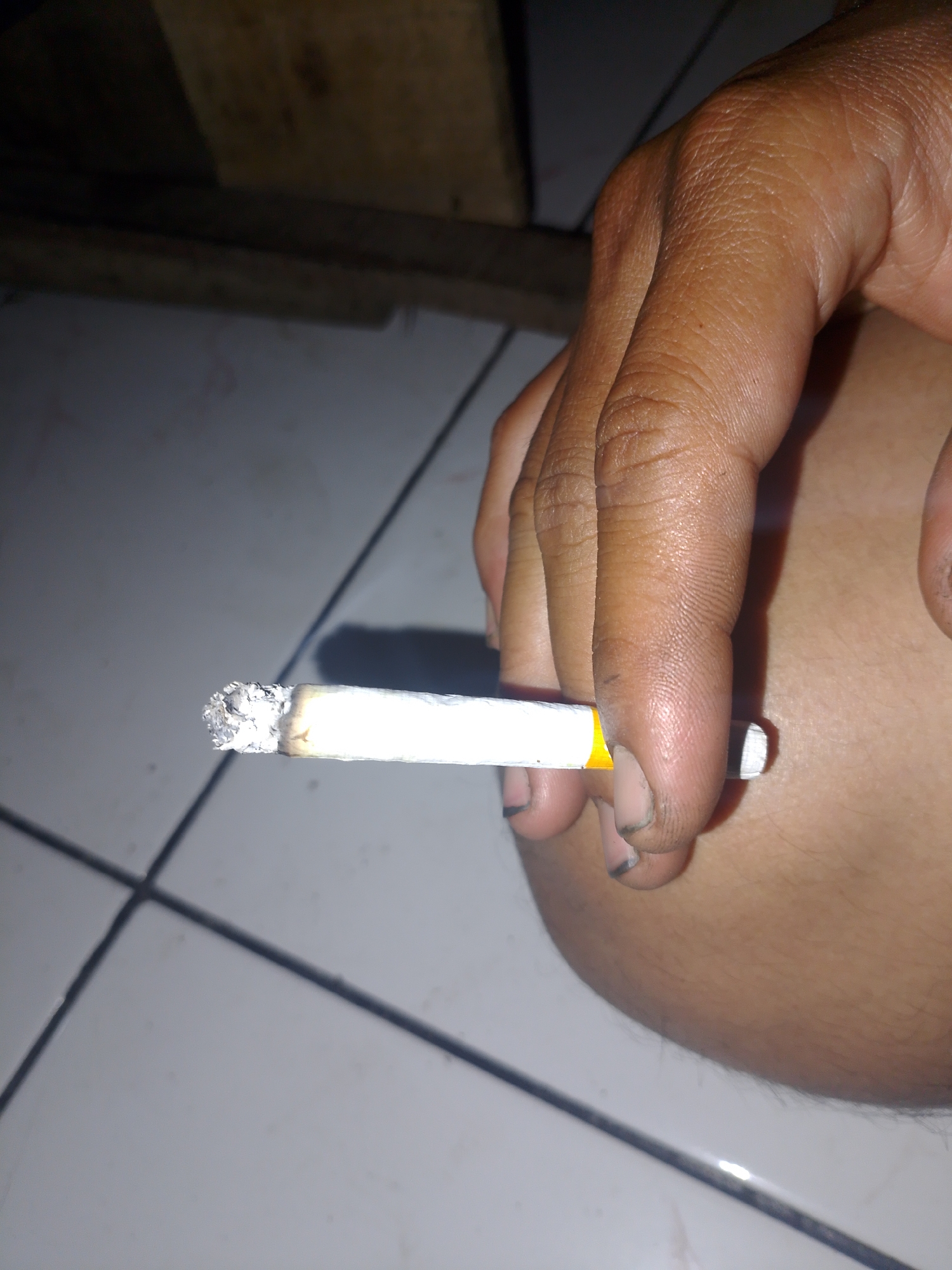 A close-up, low-angle, indoor shot focuses on a person's hand holding a lit cigarette. The hand, with visible skin texture and some dirt under the fingernails, grips the cigarette between the thumb and index finger. The cigarette, held horizontally, shows a glowing ember at its tip and a white paper wrapper with a yellow band near the filter. The background consists of a tiled floor with visible grout lines and a dark, wooden surface at the top. The lighting is dim, creating shadows and highlighting the cigarette and hand. The scene appears to be located in Mukapayung, Indonesia.