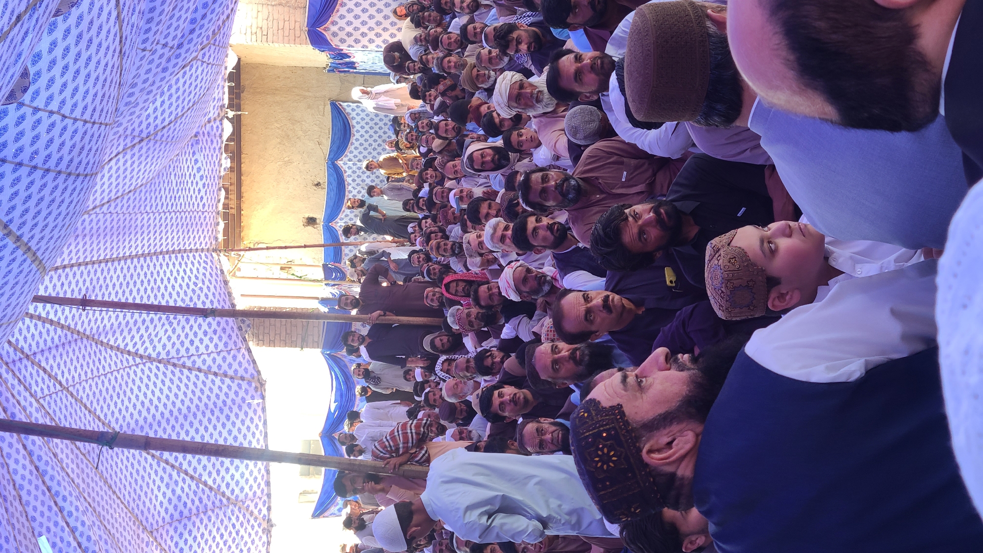 The image depicts a large gathering of men outdoors in what appears to be Panjpai, Pakistan. The men are predominantly seated and looking in the same direction, suggesting they are observing an event. Many are wearing traditional Muslim head coverings, such as turbans or prayer caps. Overhead, a tent-like structure provides shade, indicated by the blue and white patterned fabric. In the background, there is a partially visible building structure. The overall impression is of a religious or community gathering.