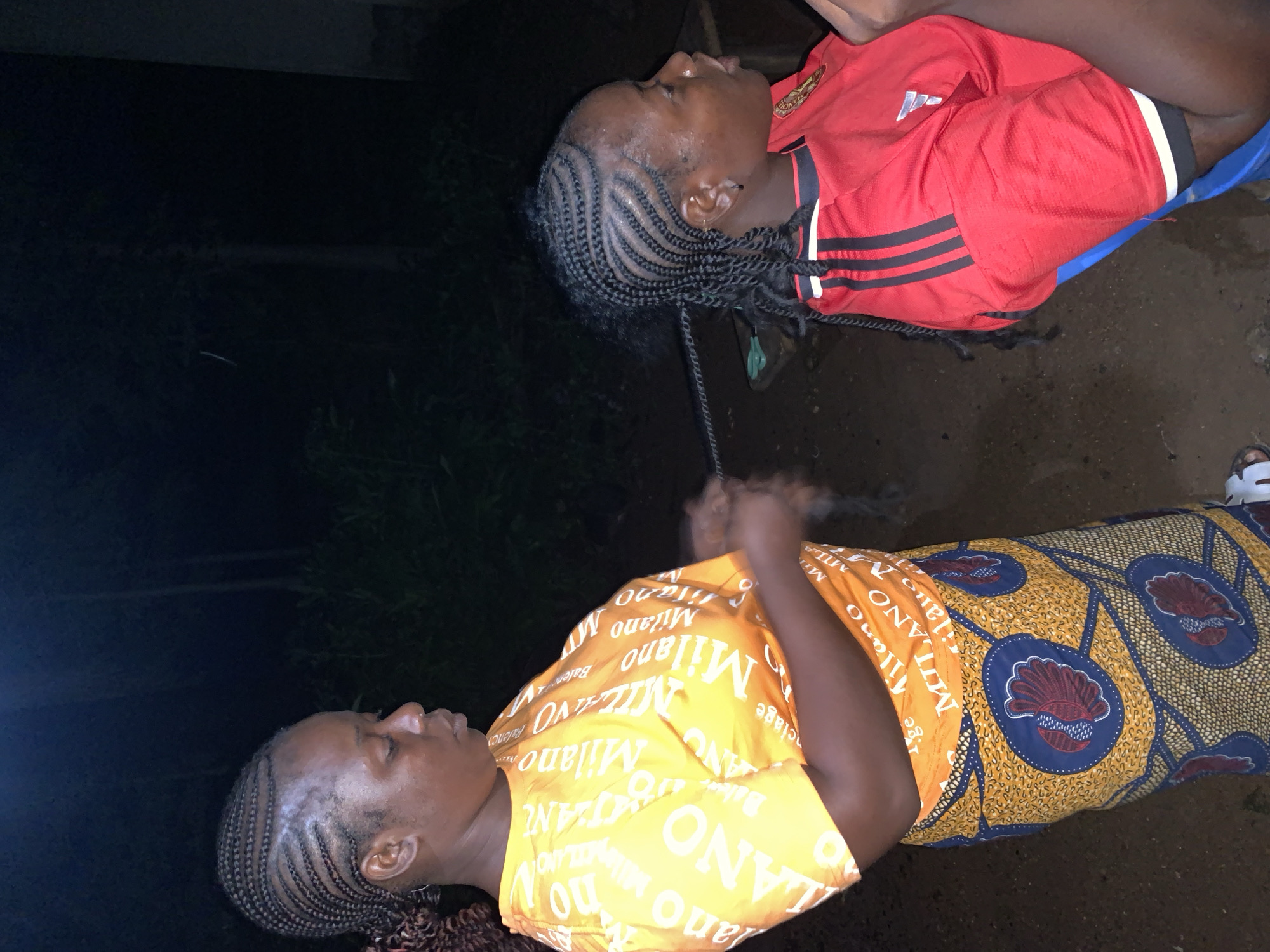 Two women are in an outdoor setting at night, possibly in Akwanga, Nigeria. The woman on the left, wearing an orange t-shirt with repeated text and a patterned skirt, is braiding the hair of the woman on the right. The woman on the right is wearing a red shirt with black and white stripes on the sleeves. The background is dark, with some suggestion of foliage and a dark structure on the right. The ground appears to be dirt. A blue chair is partially visible.