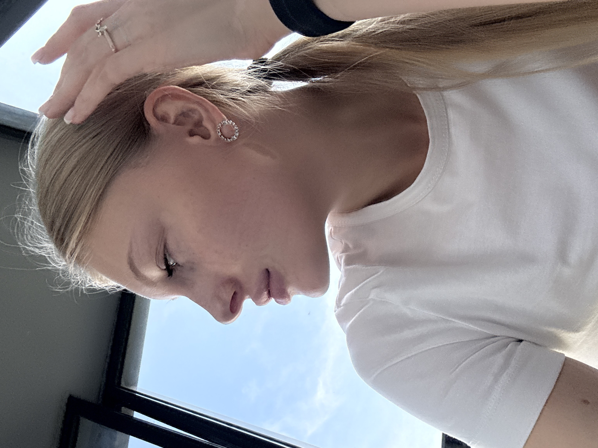 A young Caucasian woman with blonde hair pulled back into a ponytail is seen in profile, looking out a window. She is wearing a white t-shirt and has a ring on her right hand, which is touching her head. She also has a circular earring in her left ear. The background shows a bright blue sky with some clouds, indicating it is daytime. The window frame, a dark vertical line, is visible on the left side of the image. There is no indication of the city of Perm, Russia, or any other specific location within the image.