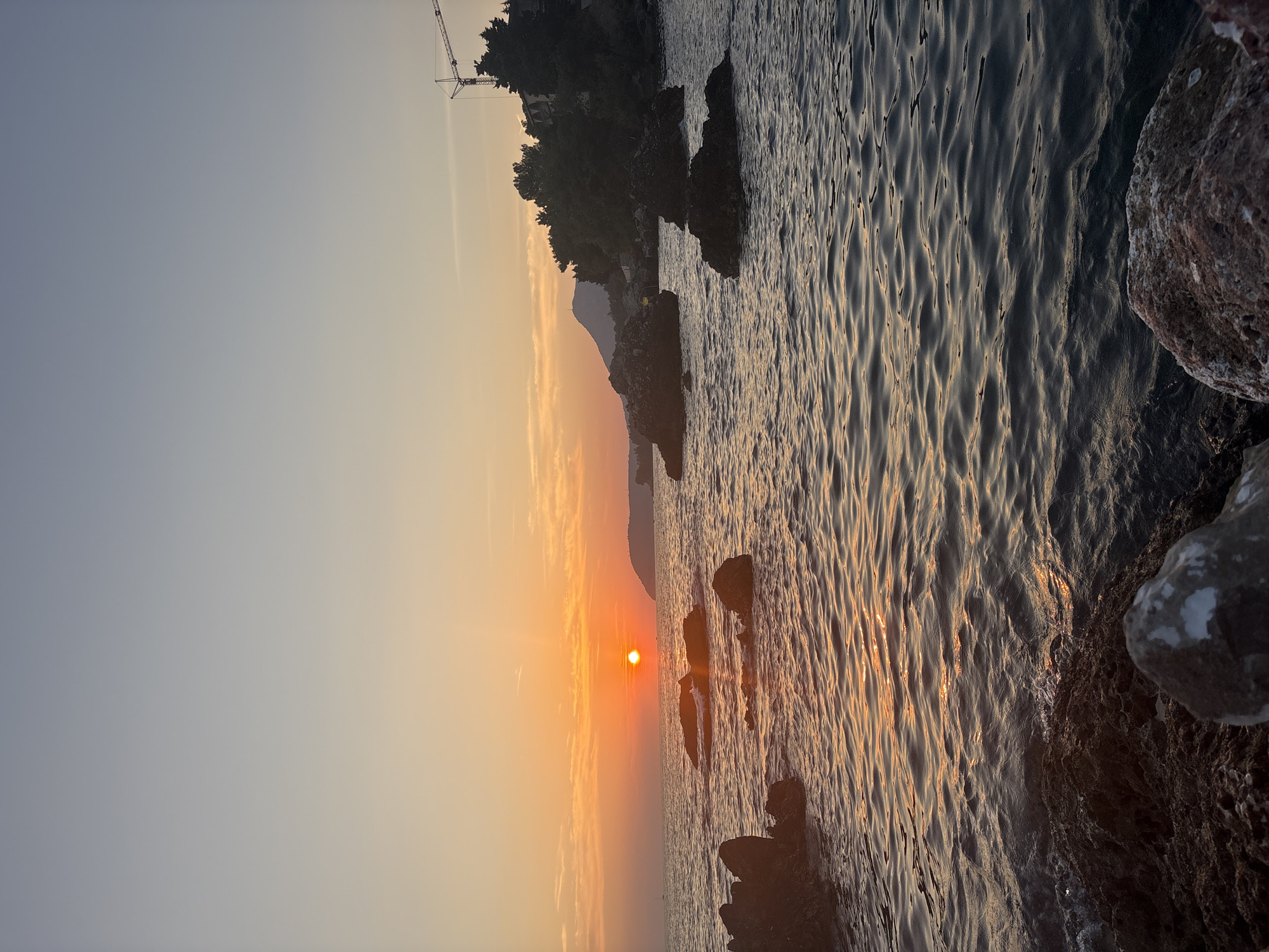 This image depicts a sunset over the Adriatic Sea, viewed from the rocky shoreline of Bar, Montenegro. The sun, a bright orange orb, is low on the horizon, casting a warm glow across the water. The sea is calm, with gentle ripples reflecting the colors of the sunset. Several dark rocks jut out of the water, some partially submerged. In the background, a forested hillside slopes down to the sea, with a few buildings and a construction crane visible amongst the trees. The sky above is a gradient of soft orange and blue, with a few wispy clouds. The overall scene is one of peaceful natural beauty, characteristic of the Montenegrin coast.