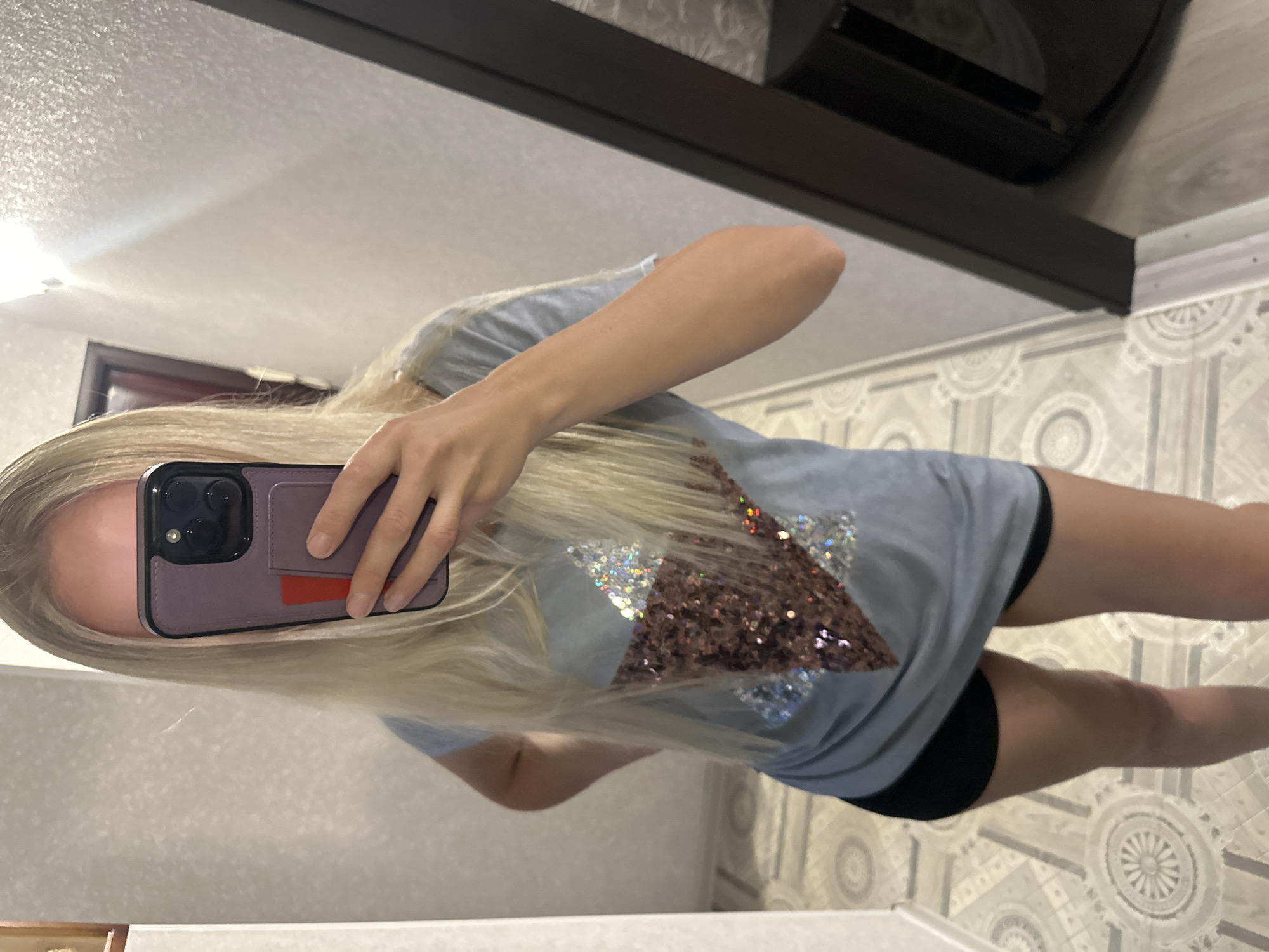 A young woman with blonde hair is taking a mirror selfie in a hallway. She is wearing a light blue t-shirt with a sequined star design and black shorts. The hallway has patterned flooring and textured walls, with a door visible in the background. A bright light fixture illuminates the space. The scene appears to be indoors, possibly in a residential setting.