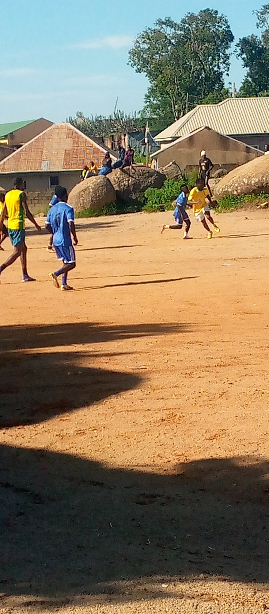 In an outdoor setting under a clear blue sky, several young males are engaged in a soccer game on a dusty, uneven field. The foreground is dominated by the sandy ground, with shadows cast from unseen objects. In the midground, four individuals are actively playing soccer. One person in a yellow and green jersey with blue shorts is running forward, looking down at the ball. Another individual in a blue jersey and shorts is also running. A third person in a blue jersey and shorts is partially visible. A fourth person in a yellow jersey is also running and appears to be dribbling the ball. In the background, a large boulder formation is present, with several people seated and standing on and around it, observing the game. Beyond the boulder, several buildings with corrugated roofs and some trees are visible, suggesting a rural or semi-urban environment. The overall impression is of an informal soccer match taking place in a community space.