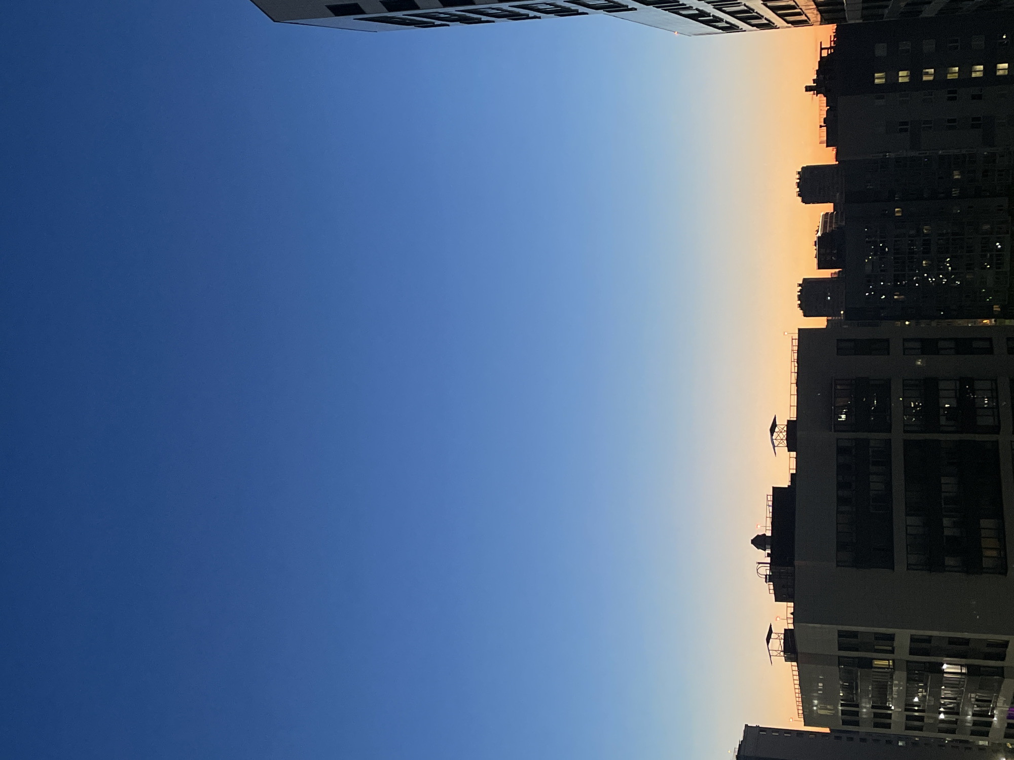 An aerial view captures the silhouette of buildings in Sinyavino, Russia, against a twilight sky. The sky transitions from a deep blue at the top to a soft orange hue near the horizon. Several multi-story buildings dominate the lower half of the image, their dark facades contrasting with the illuminated windows that dot their surfaces. Rooftops are visible, some featuring subtle architectural details like solar panels or ventilation units. The scene depicts a calm urban environment at dusk.