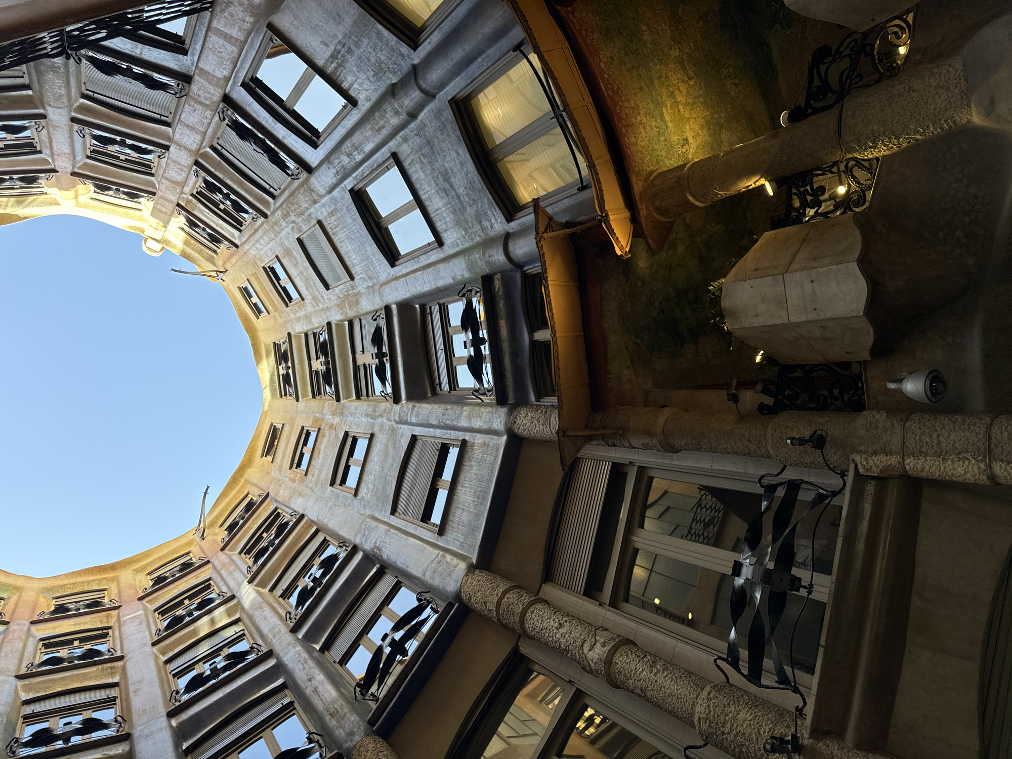 The image captures an upward view into the courtyard of Casa Milà (La Pedrera), a modernist building designed by Antoni Gaudí in Barcelona, Spain. The building’s undulating stone facade is visible, punctuated by numerous windows with ornate metalwork balconies. The windows vary in size and shape. The sky is visible through a circular opening at the top of the building. The architectural details include textured stone columns, decorative ironwork, and light fixtures. A security camera is mounted on the wall. No people are visible.