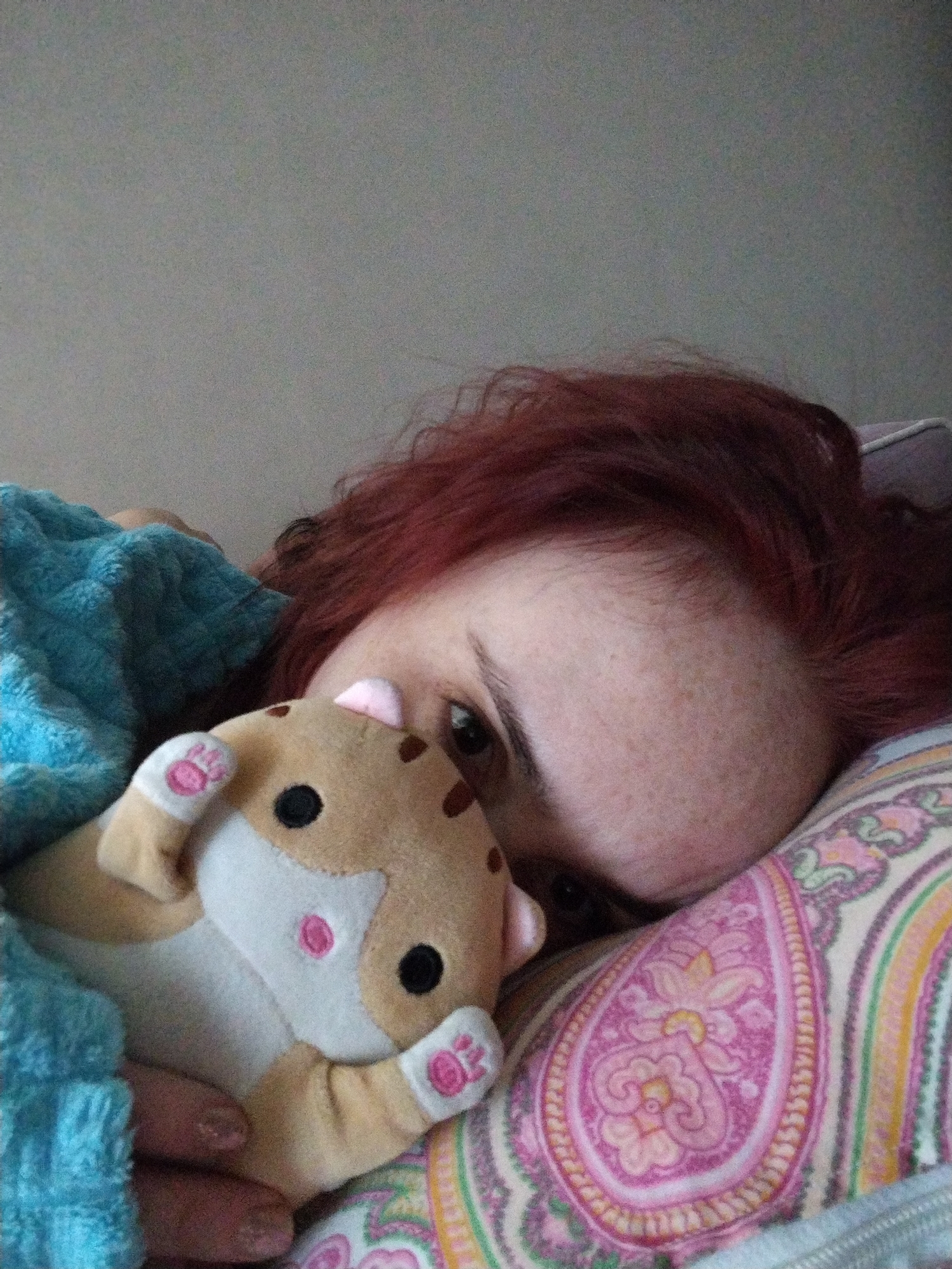 A person with red hair is lying in bed. They are on a patterned pillow, and a blue blanket is partially covering them. The person is holding a plush toy resembling a hamster, with pink details on its face and paws. The background appears to be a plain wall. There is no information in the image to indicate that it was taken in Izhevsk, Russia.