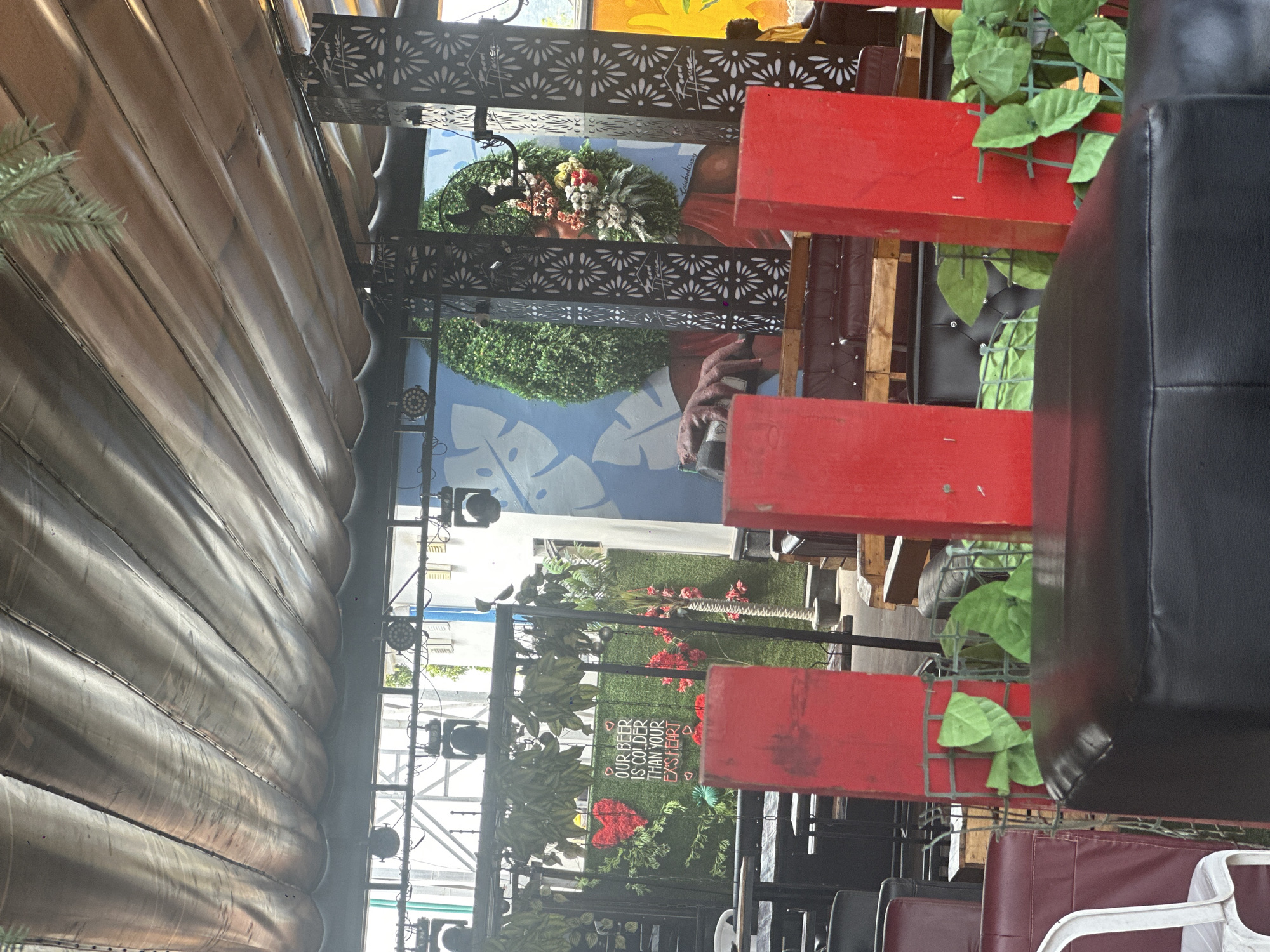 The image depicts an outdoor seating area, likely a restaurant or bar, with a covered roof constructed from dark, corrugated material. The space is decorated with artificial greenery and a mural of a woman with an elaborate hairstyle made of foliage and flowers. Red posts are prominently featured in the foreground, along with black leather seating and a white chair. A sign with the text "OUR BEER IS COLDER THAN YOUR EX'S HEART" is visible on a green wall. The overall ambiance suggests a trendy and casual establishment.