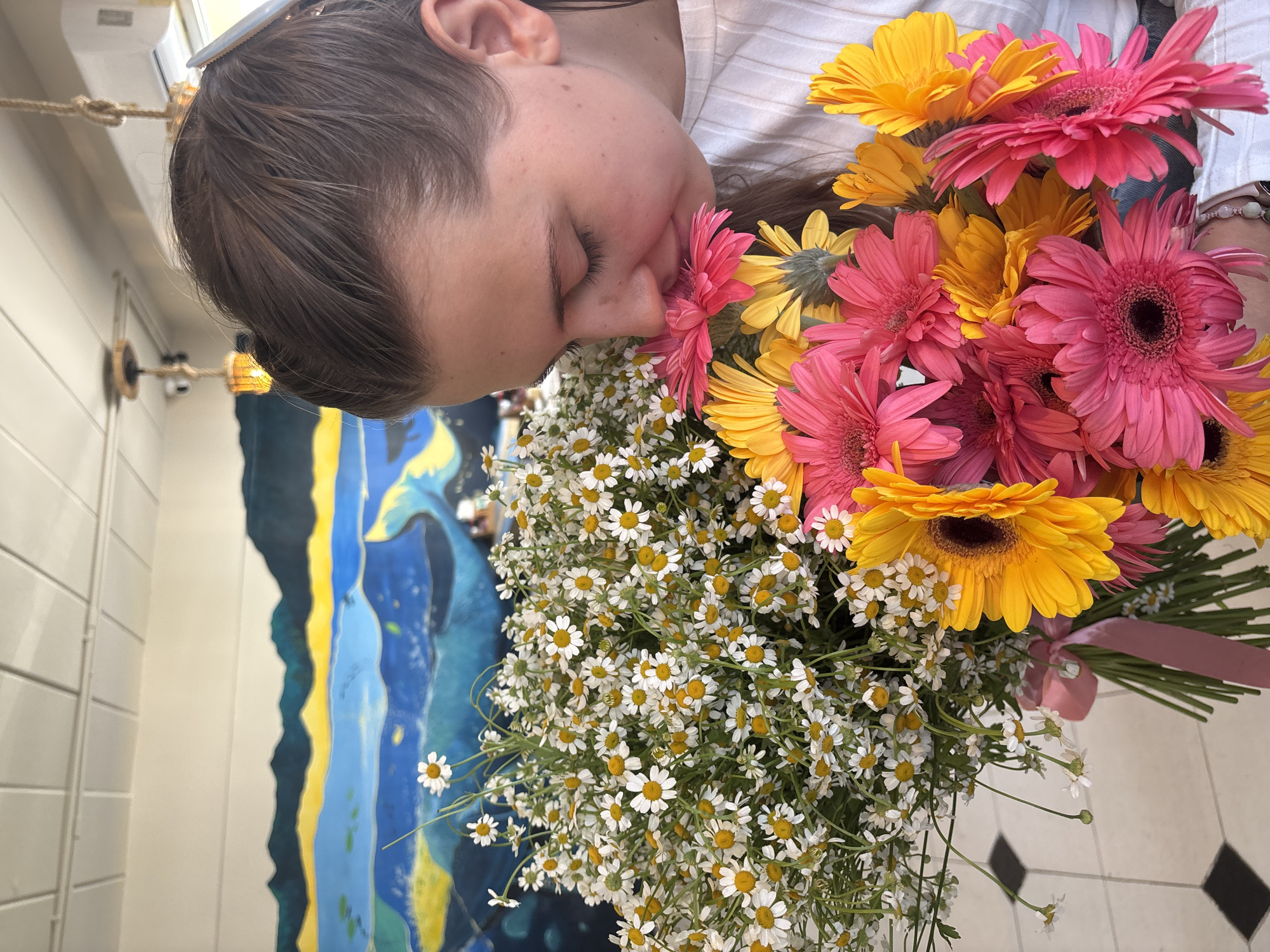 A woman is holding a large bouquet of flowers, including daisies and pink and yellow gerbera daisies. She appears to be smelling one of the flowers with her eyes closed. The scene is likely indoors, with a painting visible on the wall behind her, featuring shades of blue, yellow, and dark colors. The floor has a black and white checkerboard pattern. The location is Phuket City Municipality, Thailand.