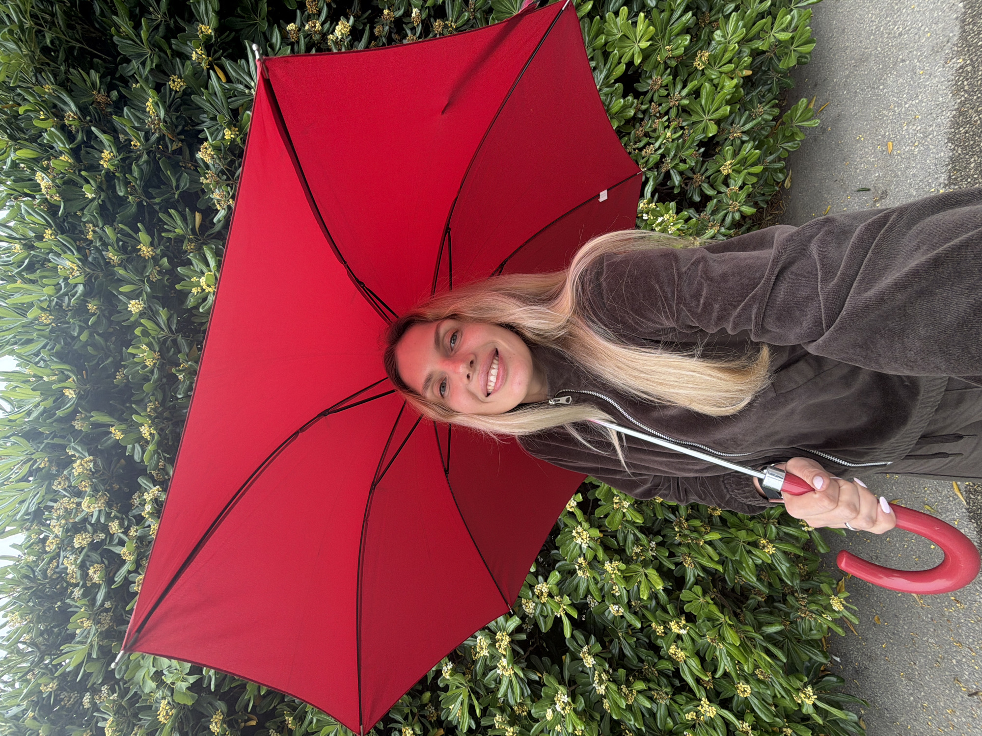 A young, light-skinned woman with blonde hair is holding a red umbrella. She is wearing a brown corduroy jacket. The background features a green bush with small yellow flowers. A sidewalk is also visible. There is no indication of the location being Budva, Montenegro.