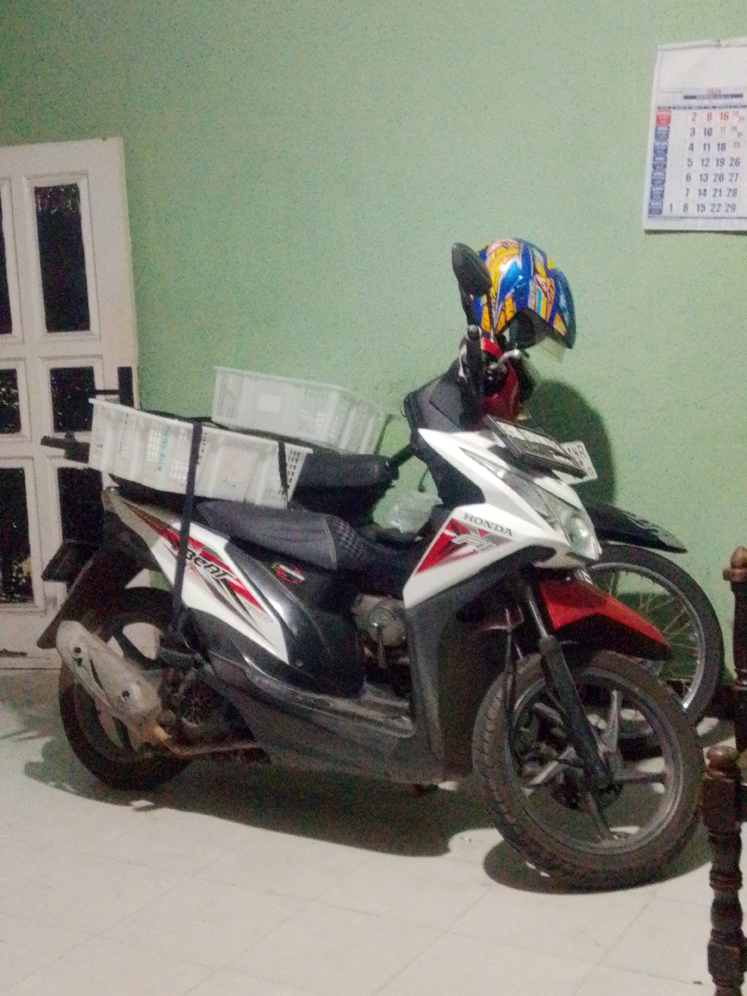 Two motorcycles are parked indoors, near a light green wall. The motorcycle in the foreground is white and red, with the "BEAT" logo visible on its side. It has two white baskets attached to its rear. The second motorcycle, partially obscured, is black with a colorful helmet placed on its handlebars. A calendar is visible on the wall in the background. The floor appears to be tiled. The scene is likely a home or storage area in Bandar Lampung, Indonesia.