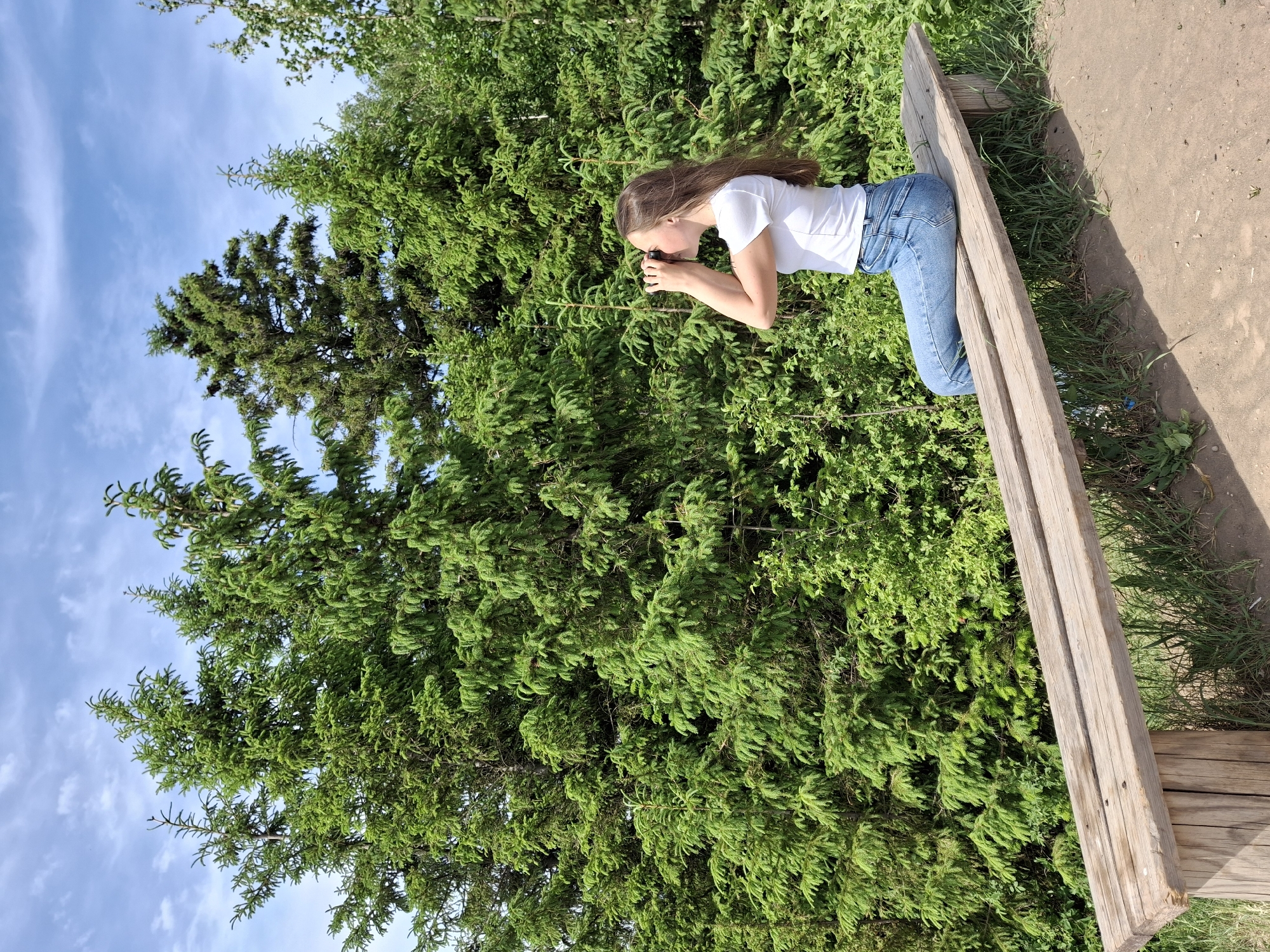 A young female with long brown hair, wearing a white T-shirt and blue jeans, is kneeling on a weathered wooden structure, possibly a low fence or a bench. She is holding a small black object up to her eyes, seemingly looking through it. The background consists of dense green foliage, including trees, with a patch of blue sky visible in the upper left corner. The ground to the right of the wooden structure appears to be dirt or gravel, with some grass. The location is inferred to be Ольховка, Russia, based on the user's prompt.