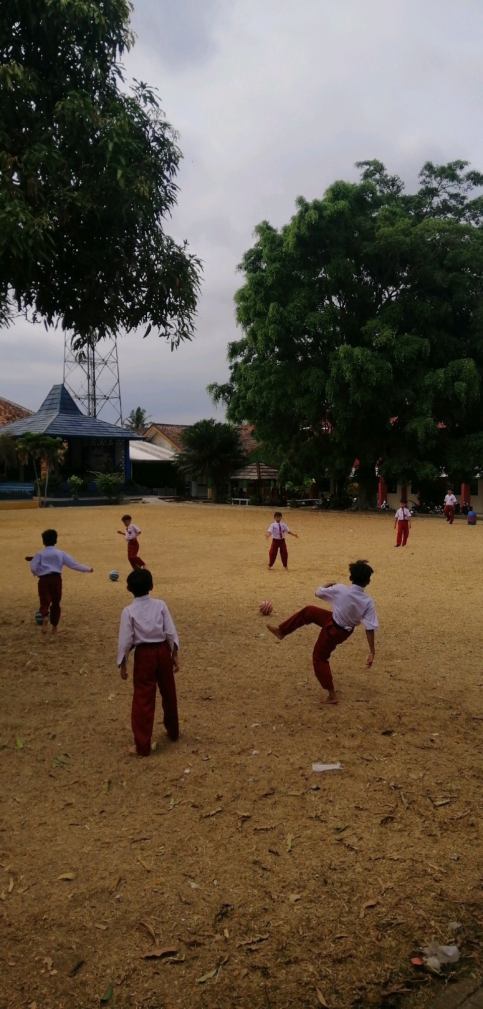 In Ajibarang, Indonesia, on a dry, sparsely vegetated field under a cloudy sky, several school-aged children in white shirts and maroon pants are engaged in a game of soccer. The children are barefoot. One child in the foreground is kicking a striped soccer ball. In the middle ground, other children are positioned on the field, some observing, others running. In the background, school buildings with red tile roofs and blue accents are visible, along with a large transmission tower and mature trees. The overall scene depicts a casual outdoor activity at what appears to be a school or community space.