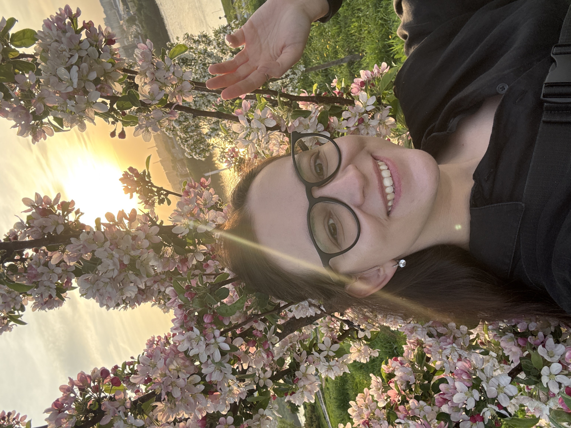 A smiling woman wearing glasses is lying in a field of flowering trees, likely crabapples, with the setting sun visible in the background. The woman is wearing a black shirt and what appears to be a backpack strap. The location is Nizhny Novgorod, Russia, suggested by the architecture visible across the river in the background. The woman's hand is raised slightly. The light suggests the photo was taken near dusk.