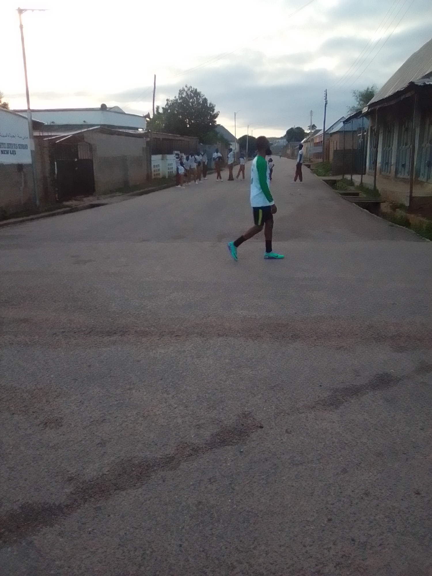 In Liji Hill, Nigeria, an outdoor scene unfolds at dusk. A group of young individuals, seemingly students, are captured mid-action on a paved road. One young person, wearing a green and white athletic outfit and blue sneakers, is prominently featured walking towards the right of the frame, with their gaze directed forward. Behind them, a line of other young people, also dressed in similar attire, are walking in procession, some facing left and others right. The background reveals a collection of buildings with corrugated metal roofs, indicative of a residential or educational area. A lamppost stands tall on the left, and utility wires stretch across the sky, suggesting a semi-urban environment. The sky is overcast, with the diffused light of dusk casting a soft glow over the scene. The road itself shows signs of wear, with some patches of discoloration. The overall atmosphere is one of calm activity, possibly a post-school event or a communal walk.