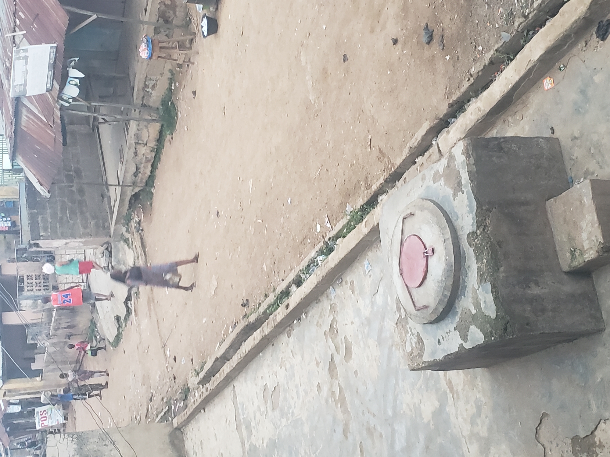 The image shows a street scene in Alalubosa, Nigeria, viewed from an elevated position. Several individuals are present on the street. One person appears to be walking alone, while others are clustered together. Some individuals are wearing brightly colored shirts. There is a concrete drainage ditch running along the edge of the street. A large, square concrete structure with a circular, red-lidded opening sits near the foreground. The surrounding buildings are made of concrete and corrugated metal. The environment seems dusty.