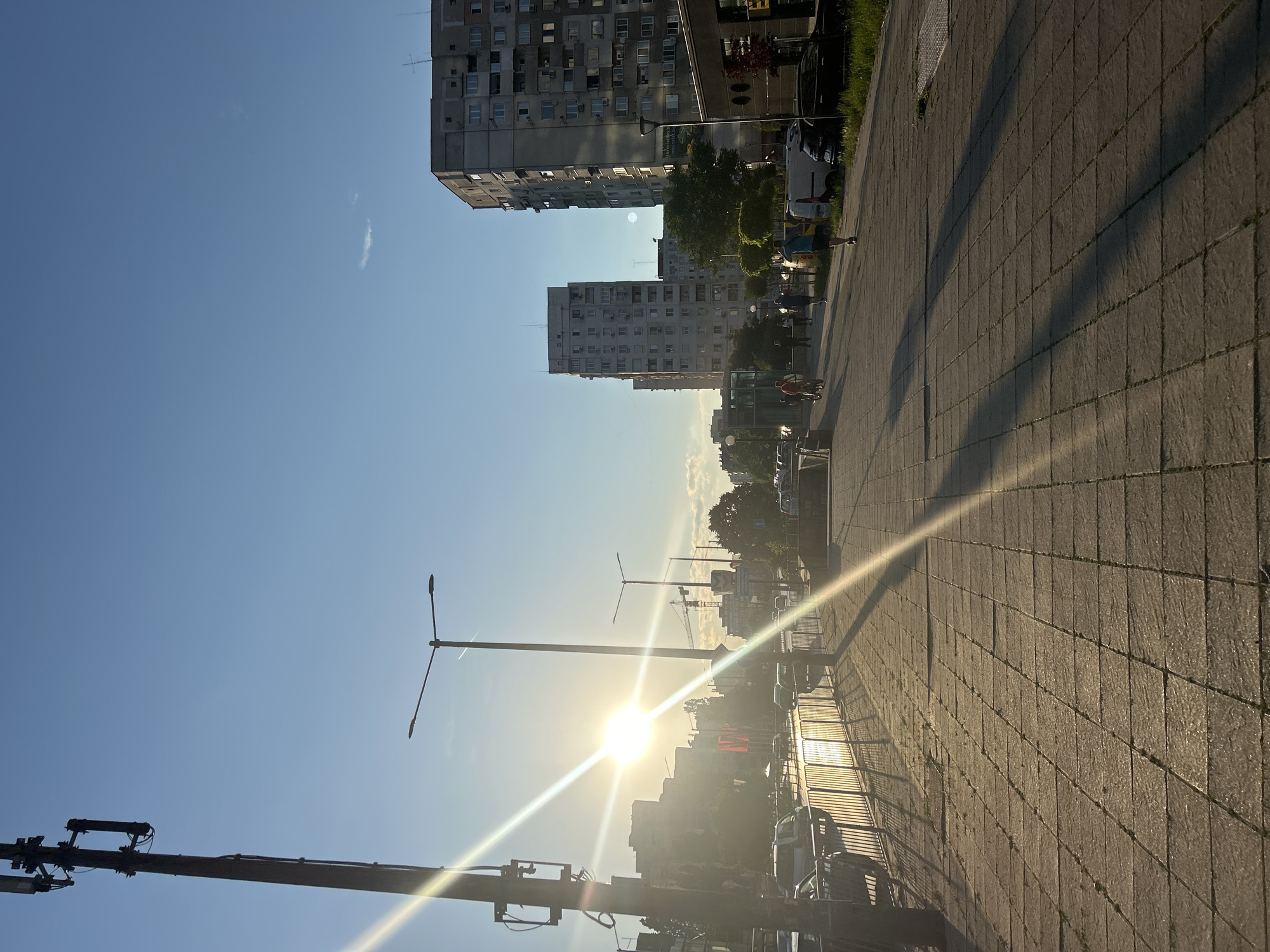The image depicts a sunlit street in Sofia, Bulgaria, likely during the late afternoon or early evening due to the low angle of the sun. The perspective is from a pedestrian sidewalk paved with large, square concrete tiles, casting long shadows.
In the foreground, a metal railing runs along the edge of the sidewalk, separating it from a street where cars are visible. The sun, positioned towards the left side of the frame, is intensely bright and creates strong lens flares and reflections on the railing and the pavement.
Further down the street, several multi-story residential buildings, characteristic of Soviet-era architecture, dominate the skyline. These buildings are a mix of light and dark gray concrete, with numerous windows visible. In the background, a construction crane can be seen, suggesting ongoing development.
A few people are scattered along the sidewalk and street. Some are walking, while others appear to be cycling. A man in a blue t-shirt is seen running away from the camera. The sky is a clear, bright blue with a few wispy clouds. A cell tower is prominently positioned on the left side of the image, with its antennas silhouetted against the sky. Streetlights with a modern design are also visible along the sidewalk. The overall impression is one of urban life bathed in the warm glow of the setting sun.
